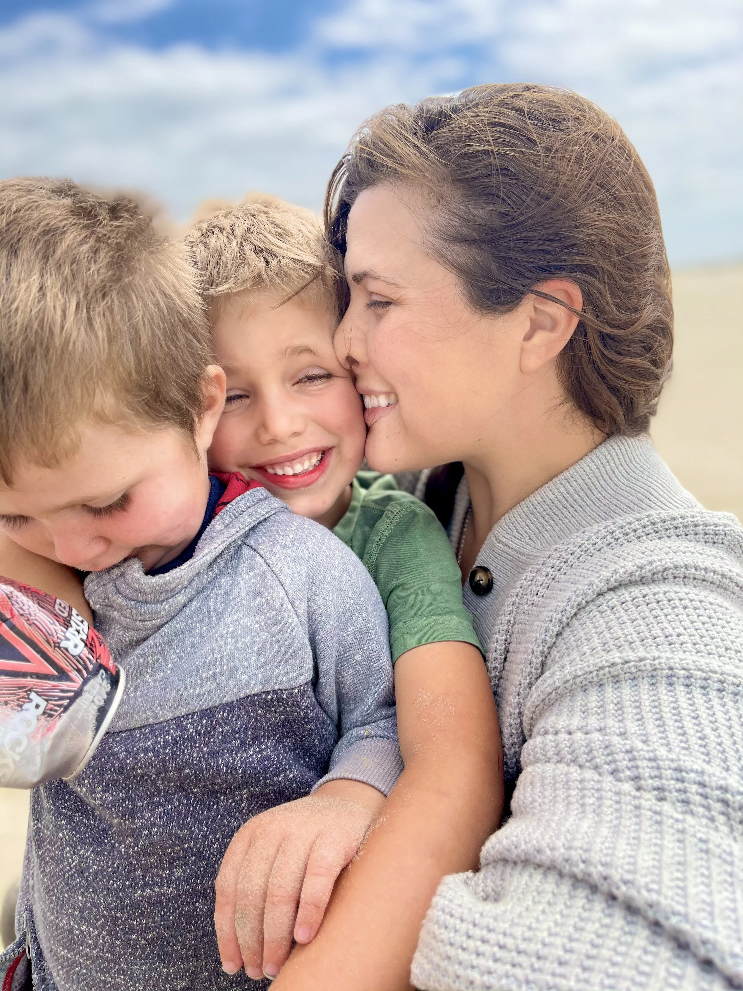 A woman embracing two young boys, giving a kiss on the cheek to one of them, on a beach with a cloudy sky in the background.