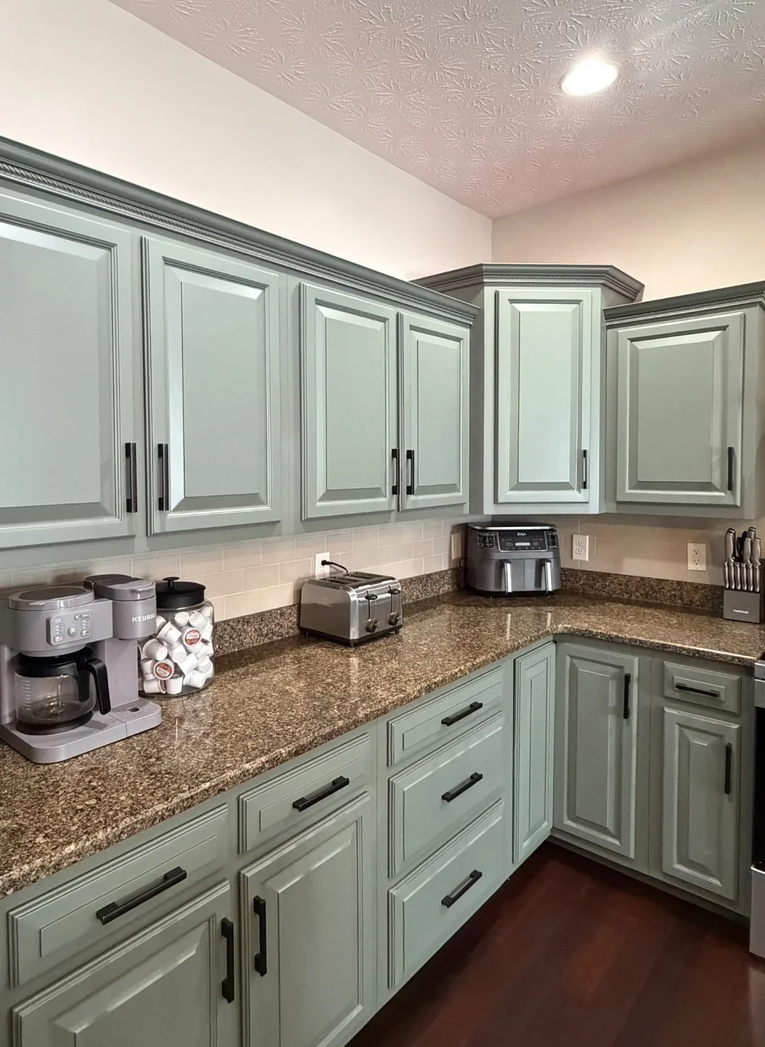 Kitchen with light green cabinets, granite countertops, small appliances including a coffee maker, toaster, and air fryer, and a dark wood floor.