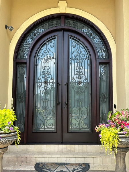 Decorative front door with intricate wrought iron design, arched window above, and surrounded by potted plants with colorful flowers.