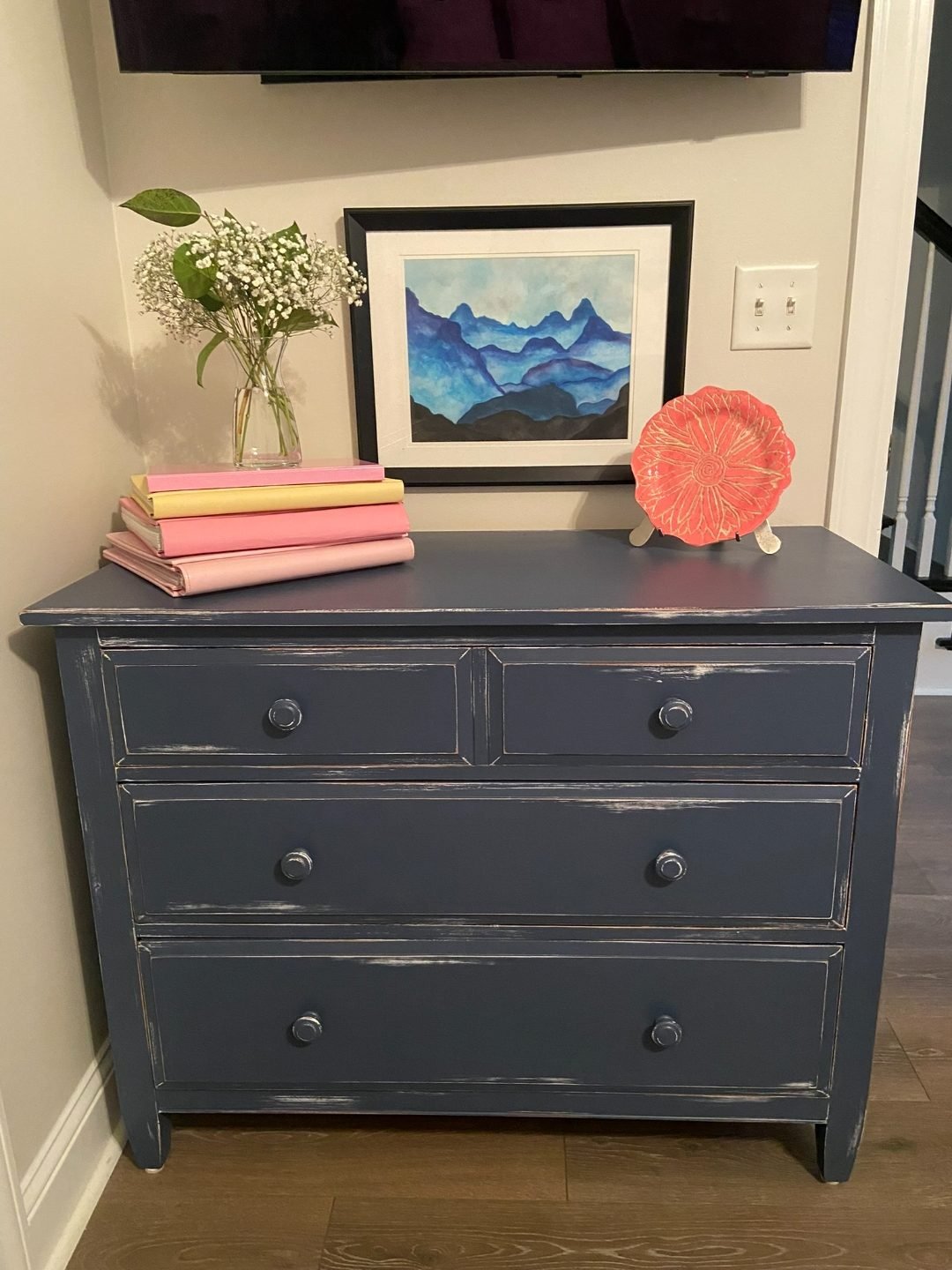 Navy/charcoal painted dresser with a lightly distressed finish and simple round knobs, styled as an entryway console with stacked books, artwork, and a small floral arrangement