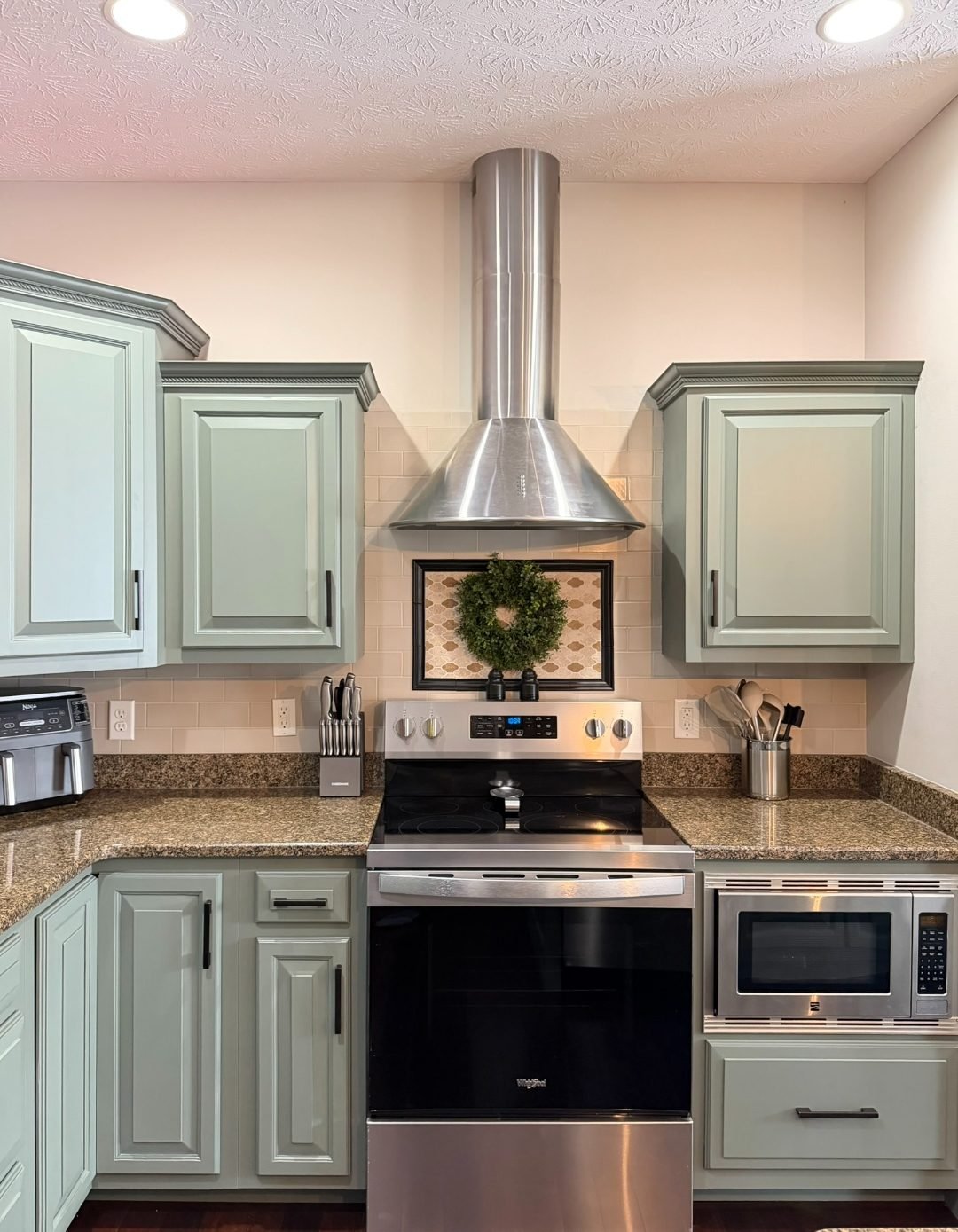 Sage green cabinet repaint with black hardware, framed by a stainless range/hood focal wall and a light subway tile backsplash.