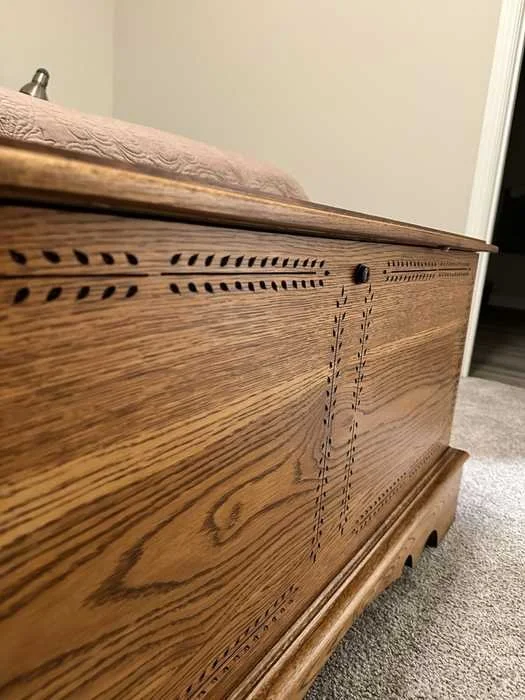 Close-up of a wooden chest with decorative carvings and a textured blanket on top, situated on a carpeted floor in a room.
