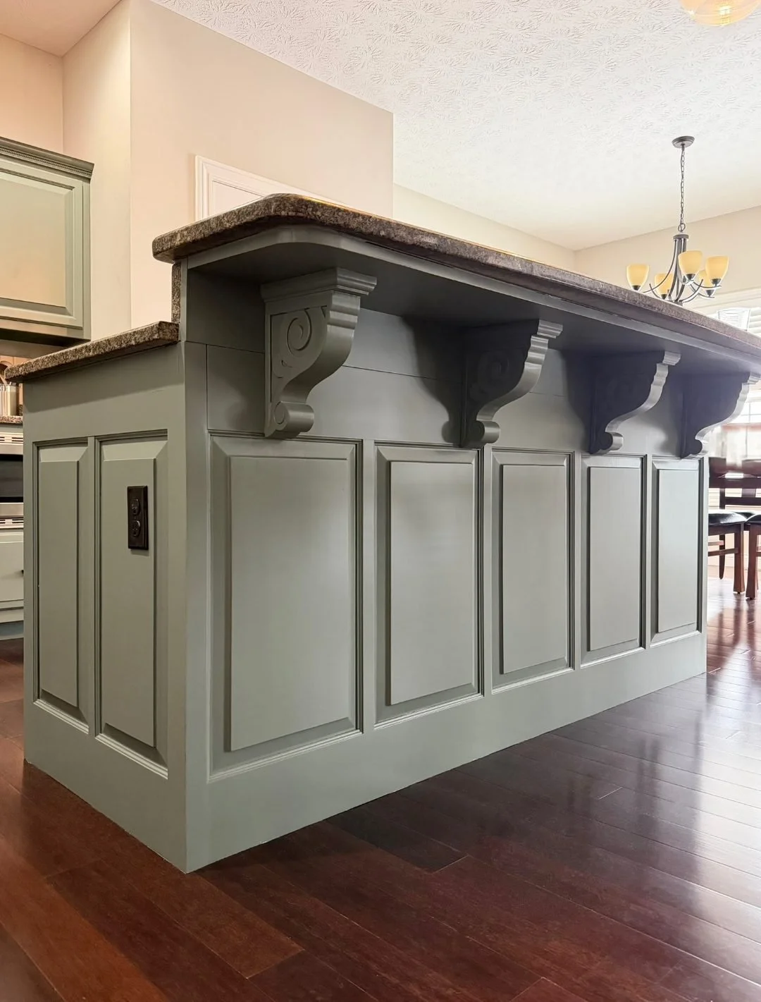 Custom-painted kitchen island in a sage green finish featuring paneled ends, decorative corbels, and a raised granite bar for seating.