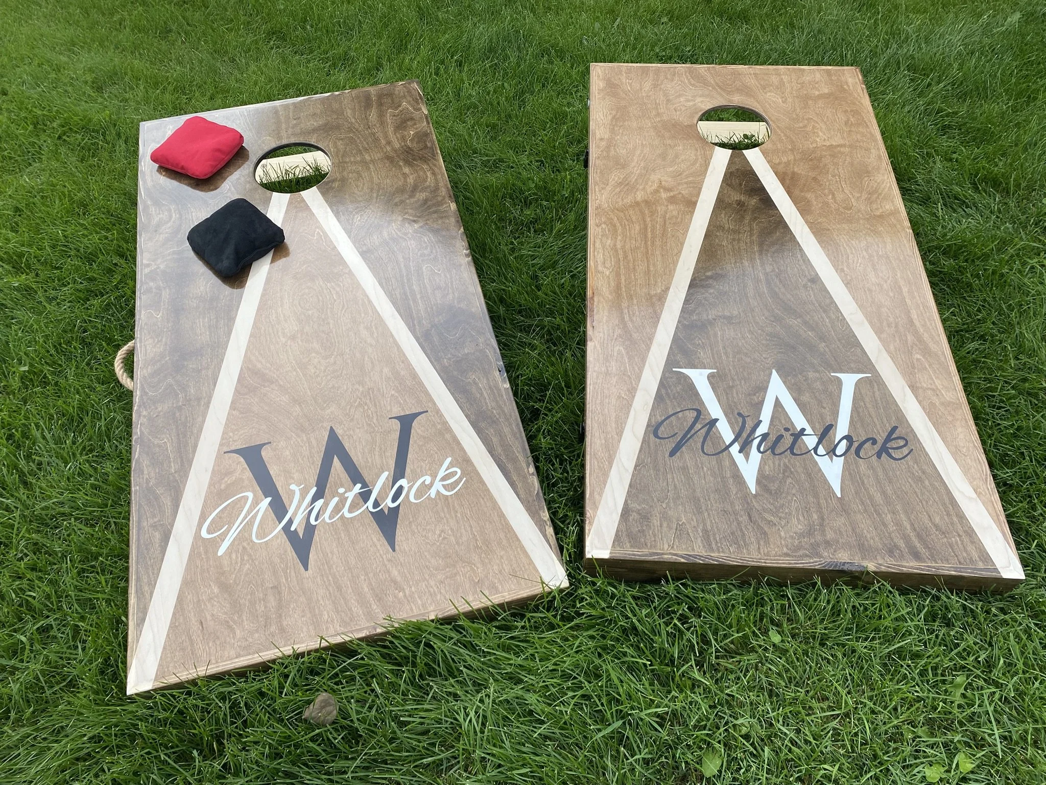 Custom Wooden cornhole game board swith a forest design at the bottom and personalized or Custom Name written across it set up on a grassy lawn with some plants and garden decorations in the background. Each board has a black and red bean bags on it.