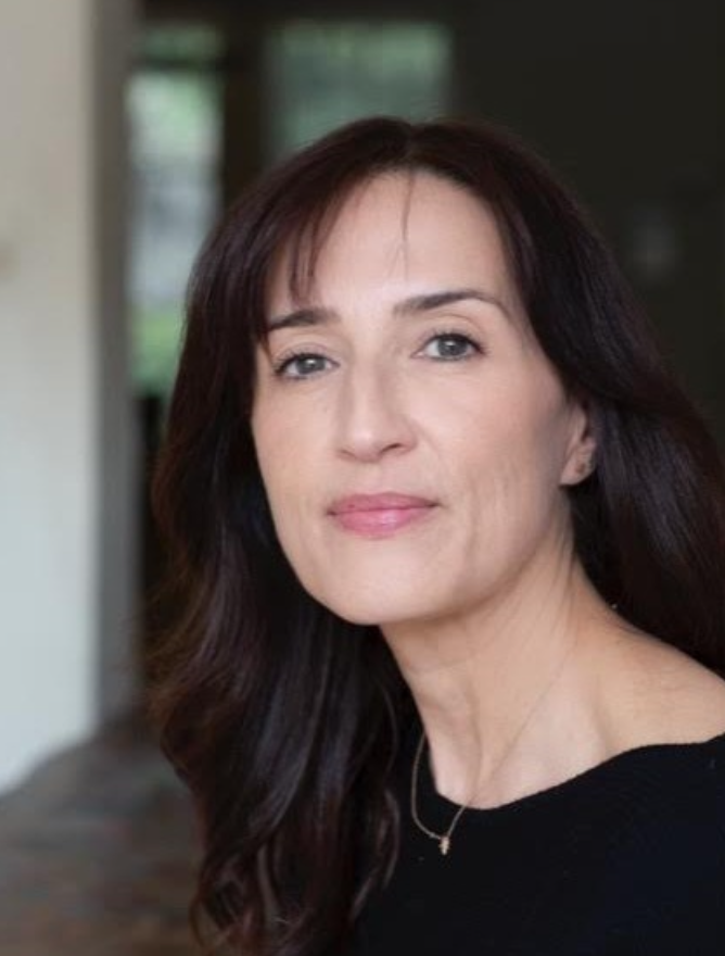 A woman with long dark brown hair and light skin looking at the camera, wearing a black top and a delicate necklace, with a blurred indoor background.