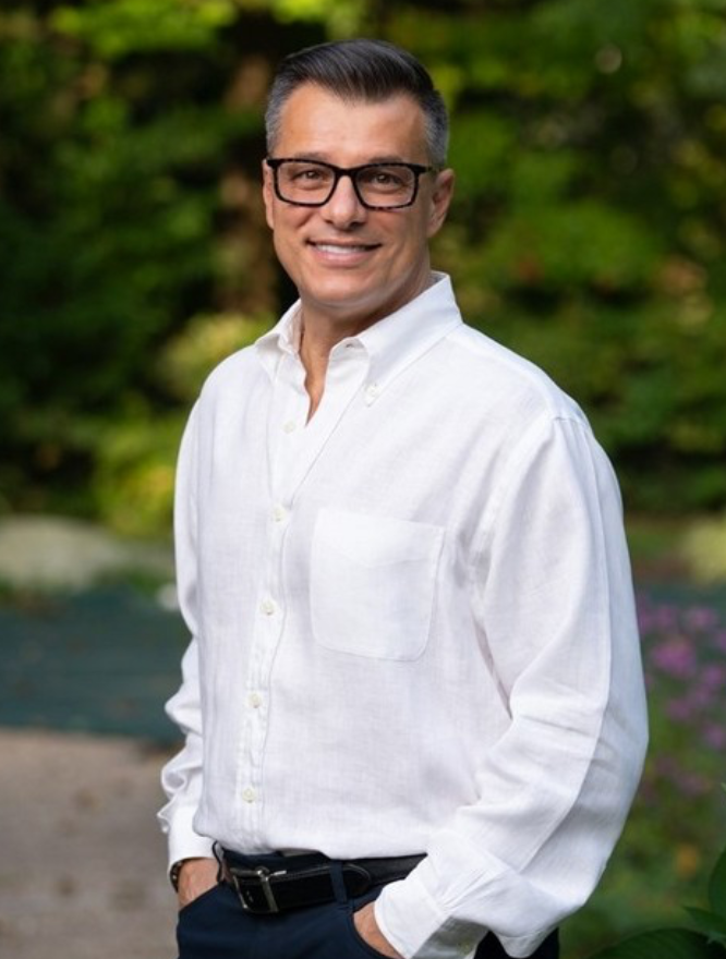 A smiling man with dark hair, wearing glasses and a white button-up shirt, standing outdoors in front of greenery.