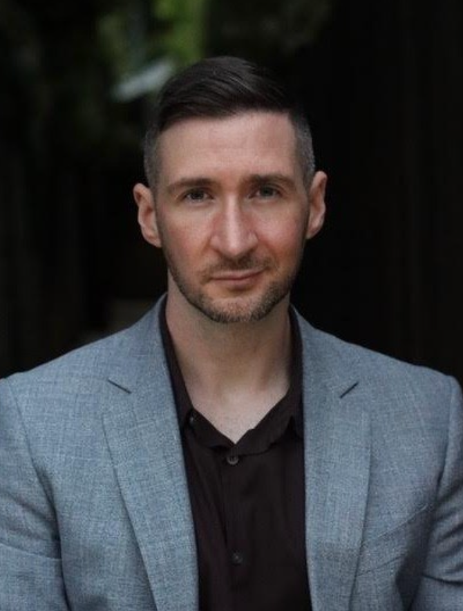 A man in a grey blazer and black shirt posing outdoors against a blurred natural background.