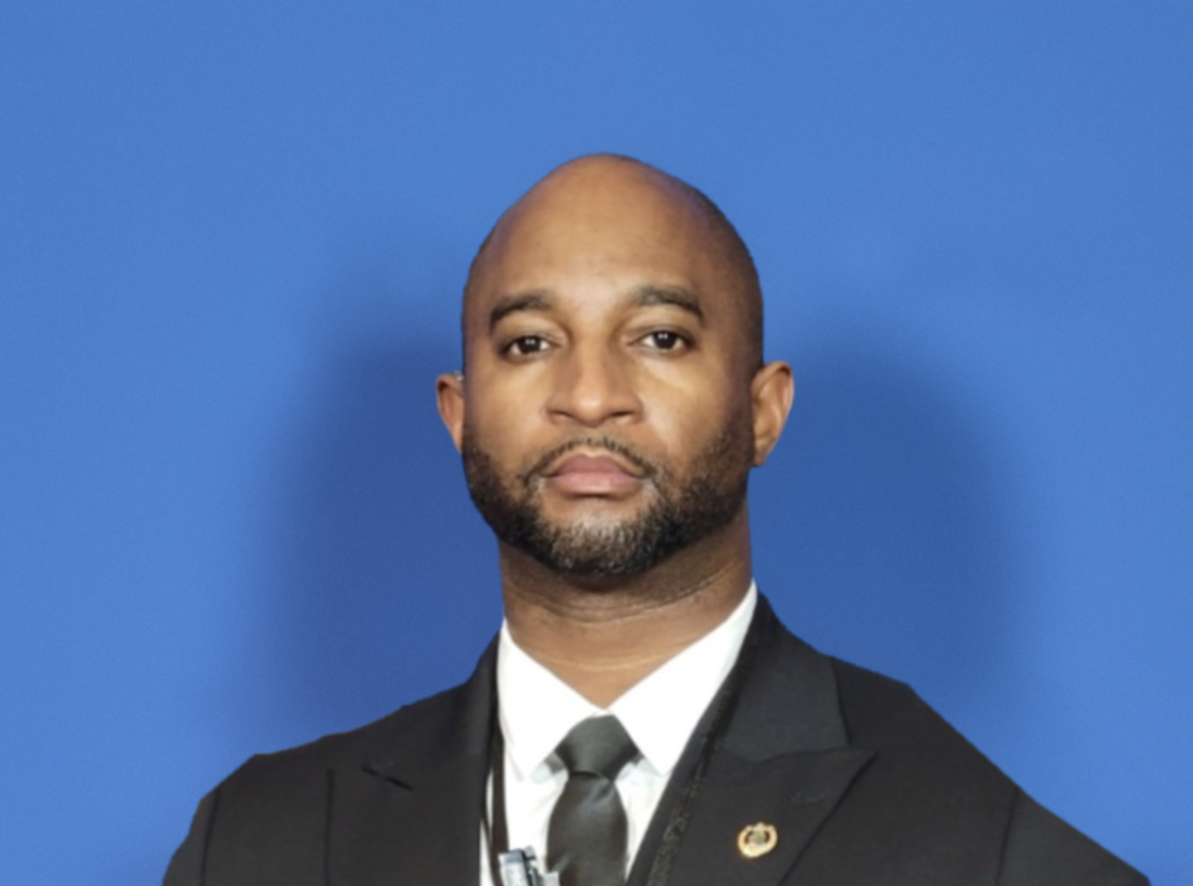 A man in formal attire, wearing a black suit with a white shirt and a black tie, standing against a solid blue background.