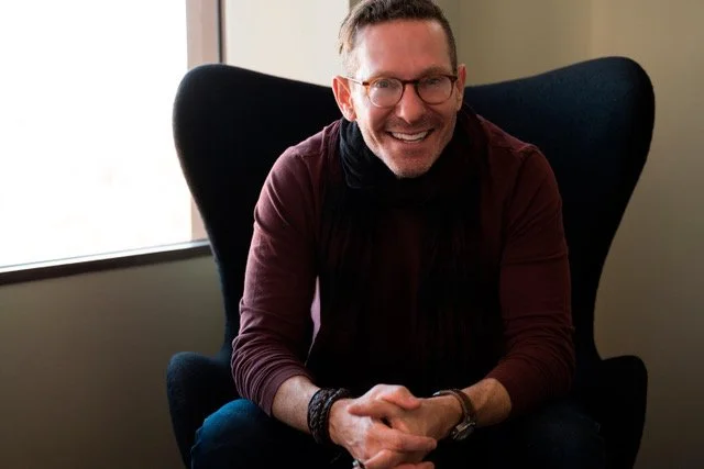 Man with glasses and a beard smiling, sitting in a black armchair near a window.