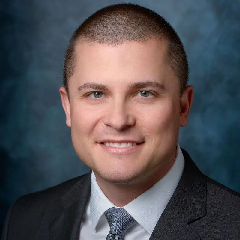 A young man with short brown hair, wearing a dark suit, white shirt, and a gray tie, smiling against a blue textured background.