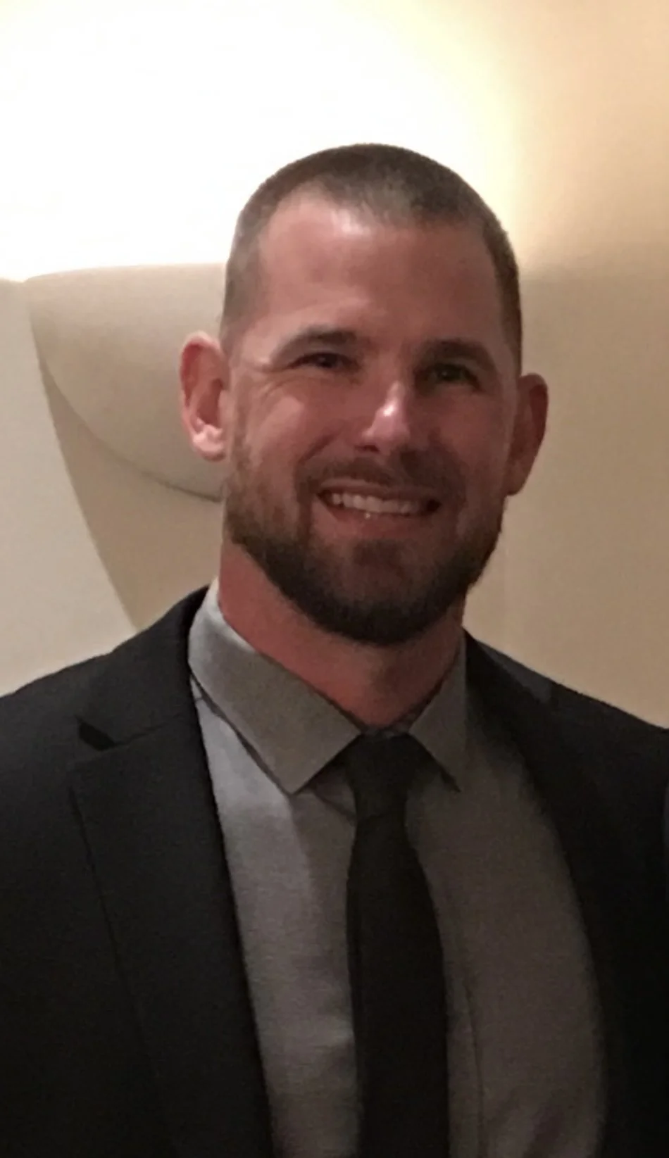 A smiling man with short hair and a beard, wearing a tuxedo with a gray shirt and black tie, standing indoors with a neutral background.