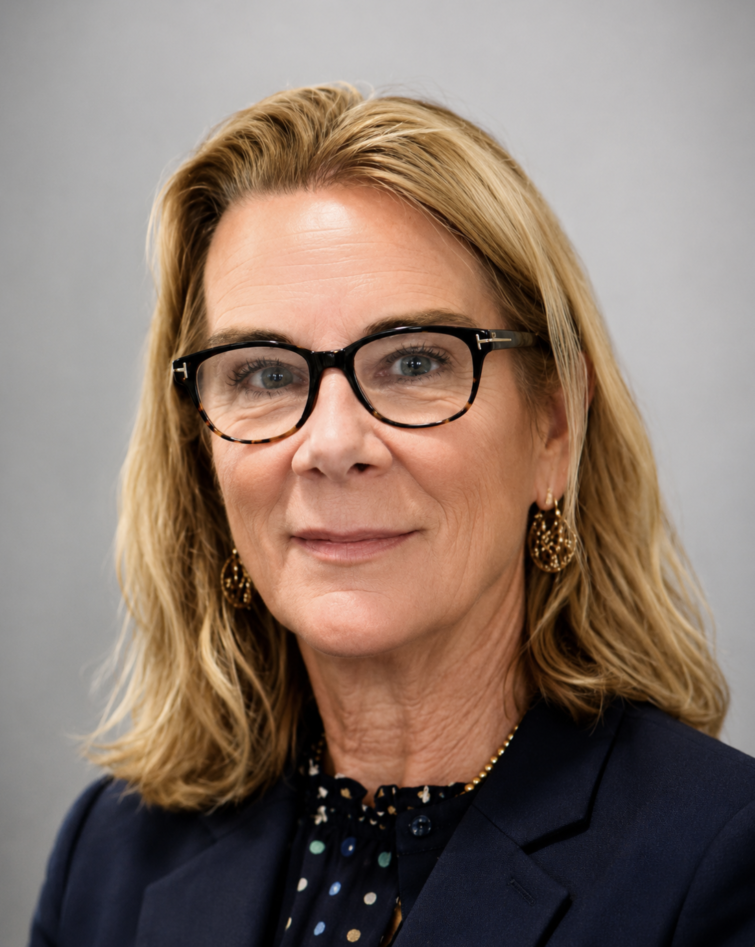 A headshot of a woman with blonde hair, blue eyes, wearing black glasses, a dark blazer, gold earrings, and a polka-dot blouse, against a gray background.