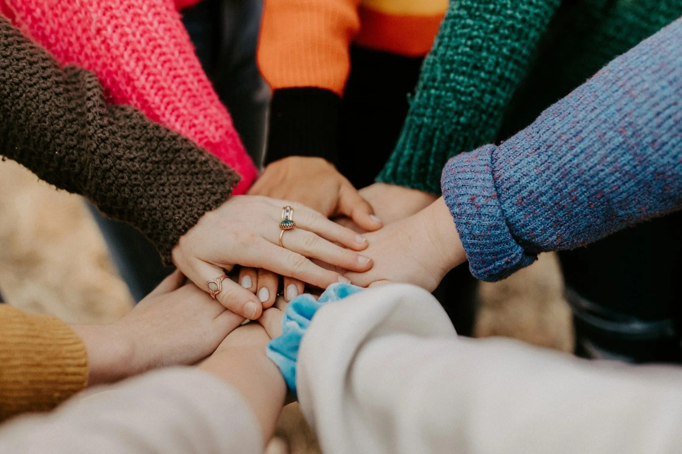 Group of people placing their hands together in a show of unity, with each person wearing a different colorful sweater.