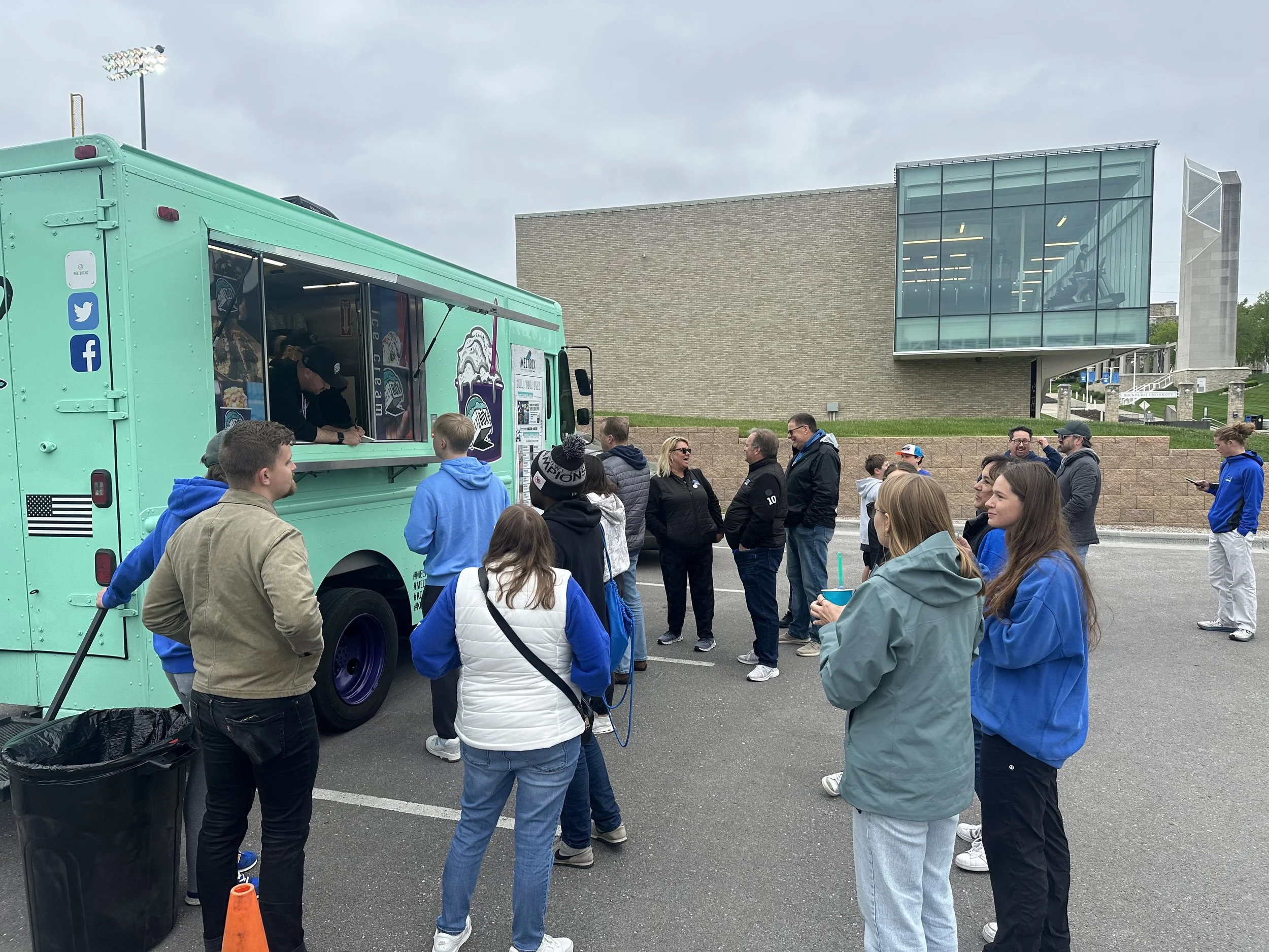 A crowd of people gathered around a teal food truck outside a modern building on a cloudy day. The food truck has social media icons on its side and a menu visible through a window. Some individuals are wearing blue jackets and appear to be waiting or talking, while others are engaged with the truck.