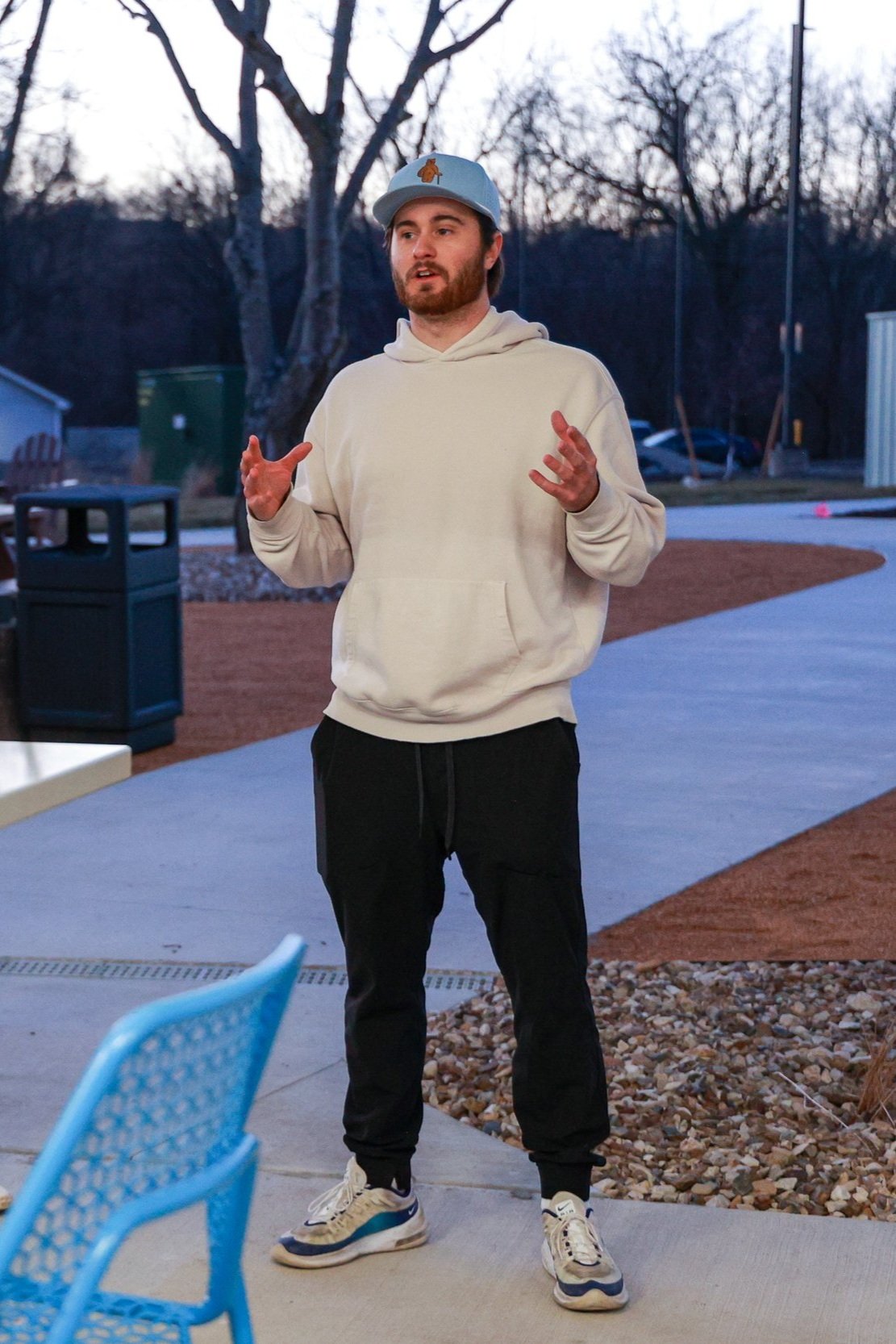 A man in a light hoodie and black pants standing outdoors, speaking with hand gestures, wearing a light blue cap with a dog logo, during twilight in a park or backyard setting.