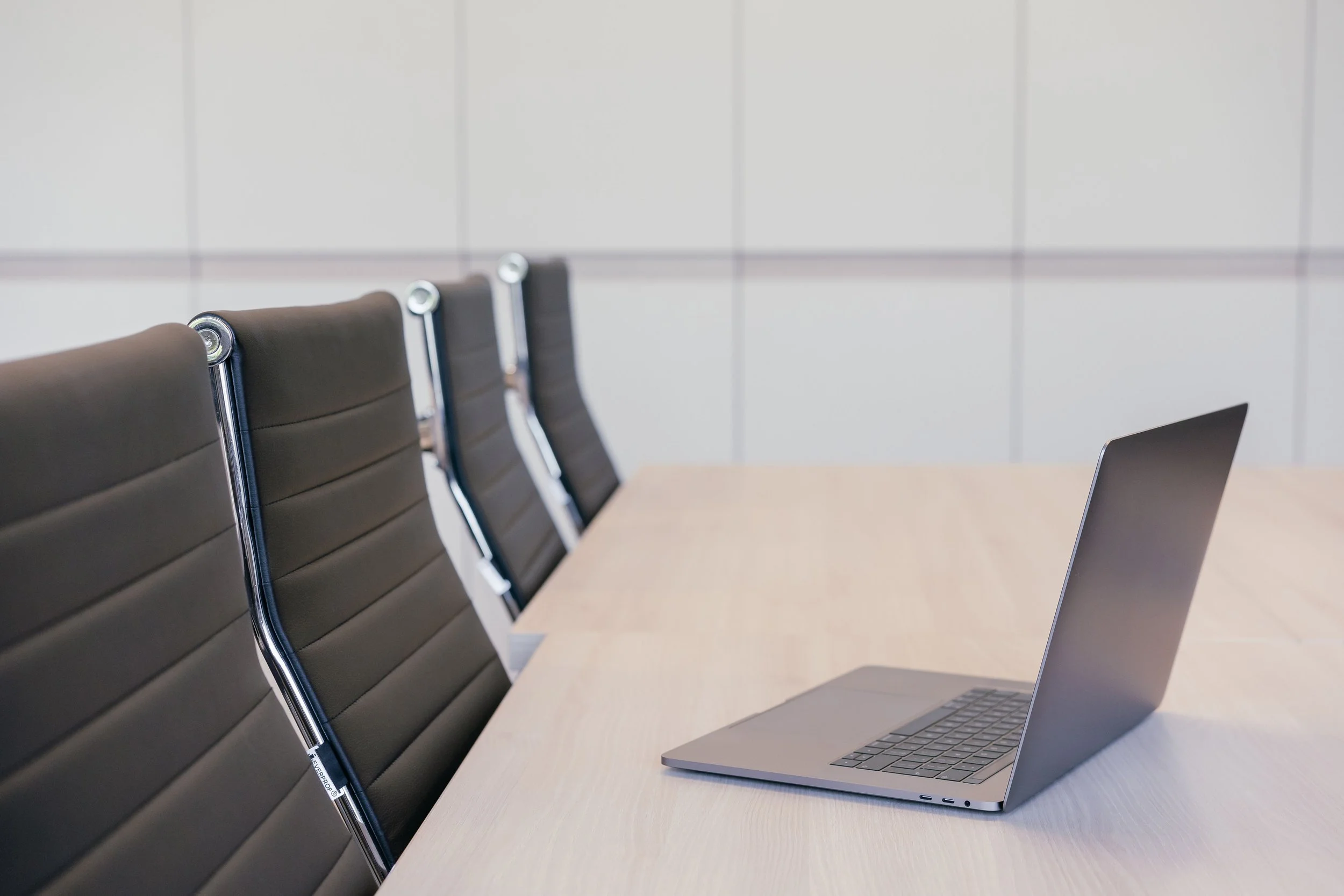 Empty conference room with four black leather chairs and a silver laptop on a wooden table.