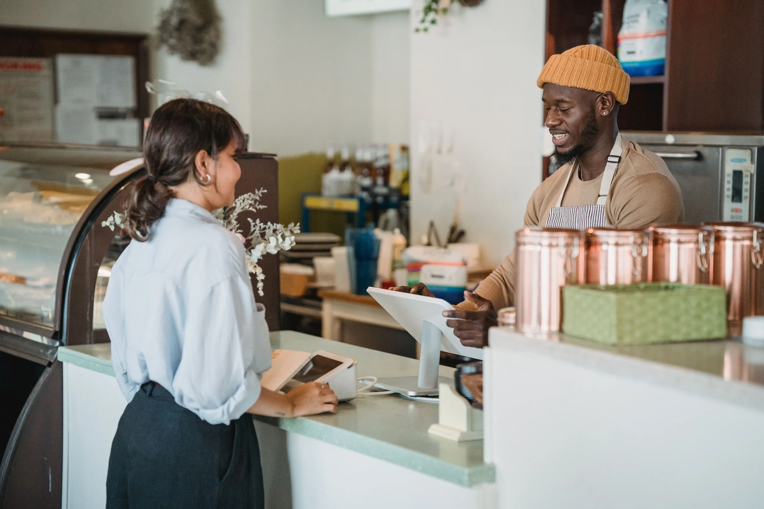 A person with dark hair in a ponytail, wearing earrings, a blue shirt, and a dark skirt, standing at a restaurant counter, facing a man with  a beard, a yellow knit cap, and a striped apron, who is showing her something on a tablet or device.