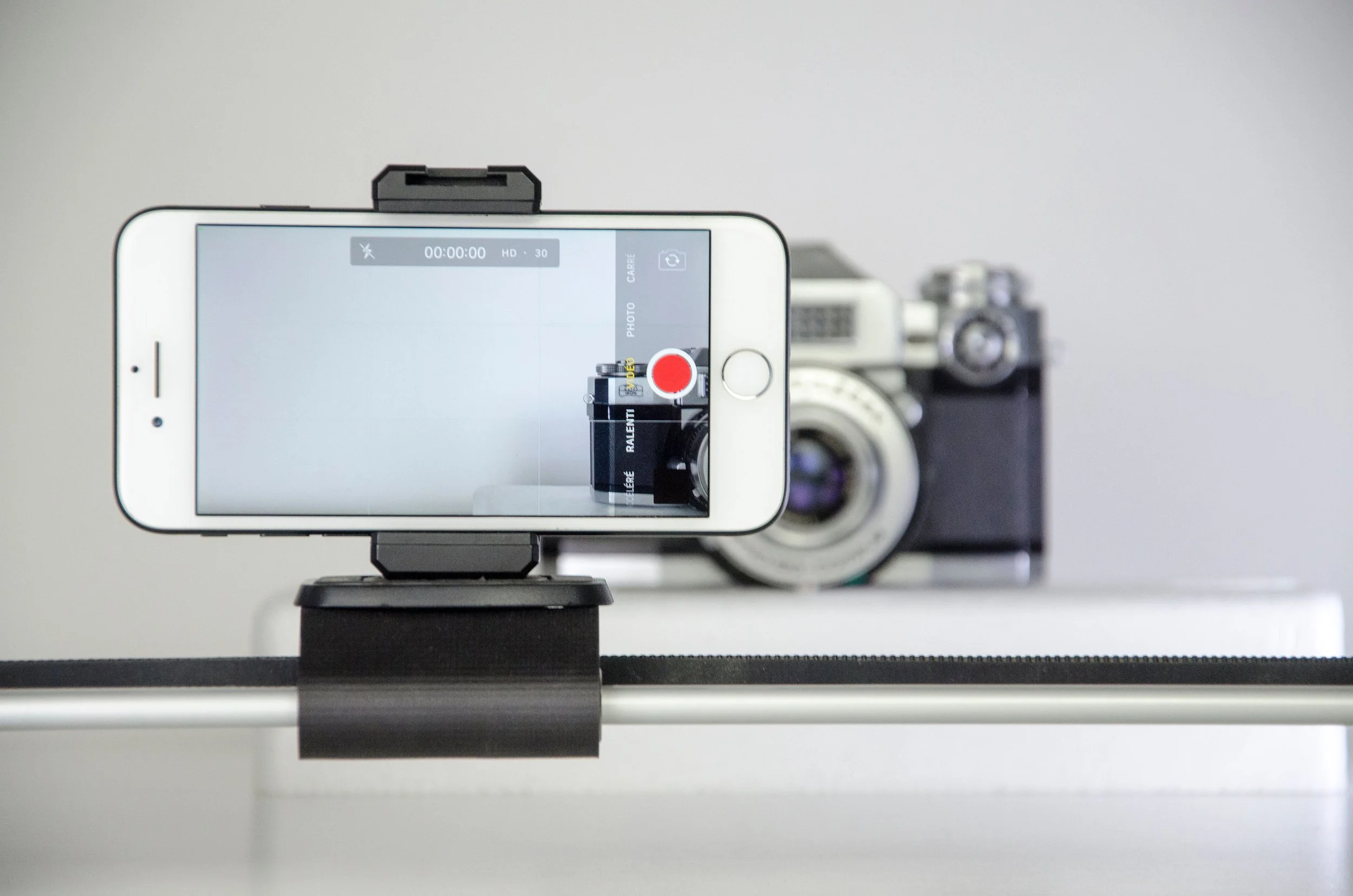 A smartphone mounted on a tripod recording a vintage camera on a white table against a plain light-colored wall.