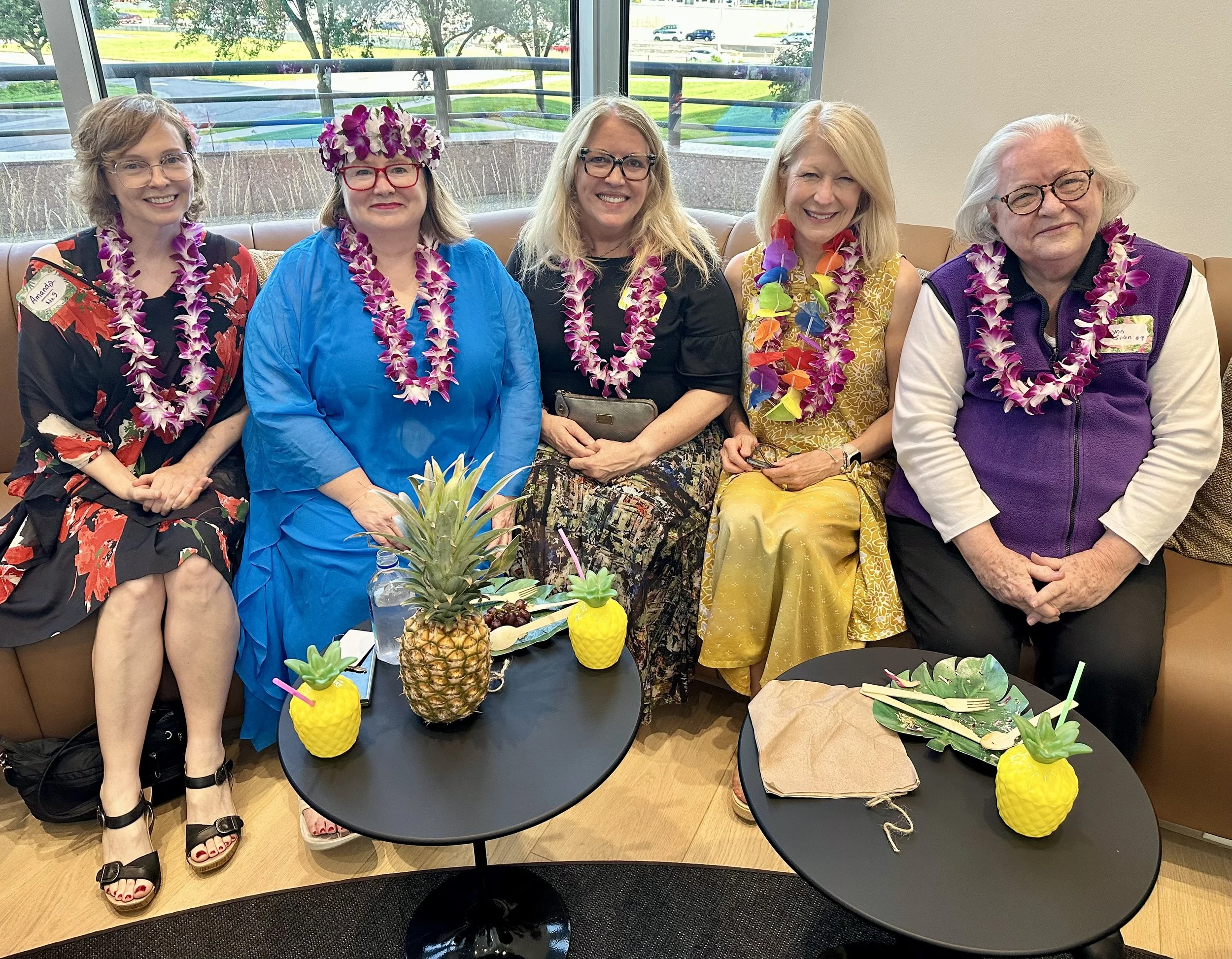 Five women sitting on a brown couch, smiling, with colorful leis around their necks. Two small tables in front of them hold pineapple-shaped cups and a pineapple. A woman on the far left wears a floral dress and black heels, the second woman from the left wears a blue dress and a floral crown, the middle woman wears a black top with a patterned skirt and glasses, the second woman from the right wears a yellow dress with a colorful lei, and the woman on the far right wears a purple vest over a white shirt and glasses.