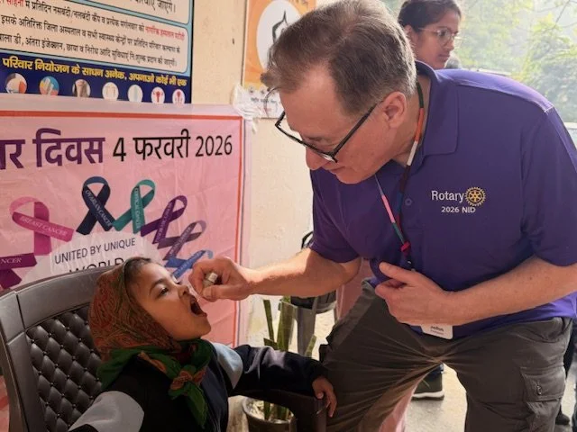 Group of people at a Rotary Club event holding plaid blankets in a wood-paneled room with a portrait and a Rotary sign in the background.