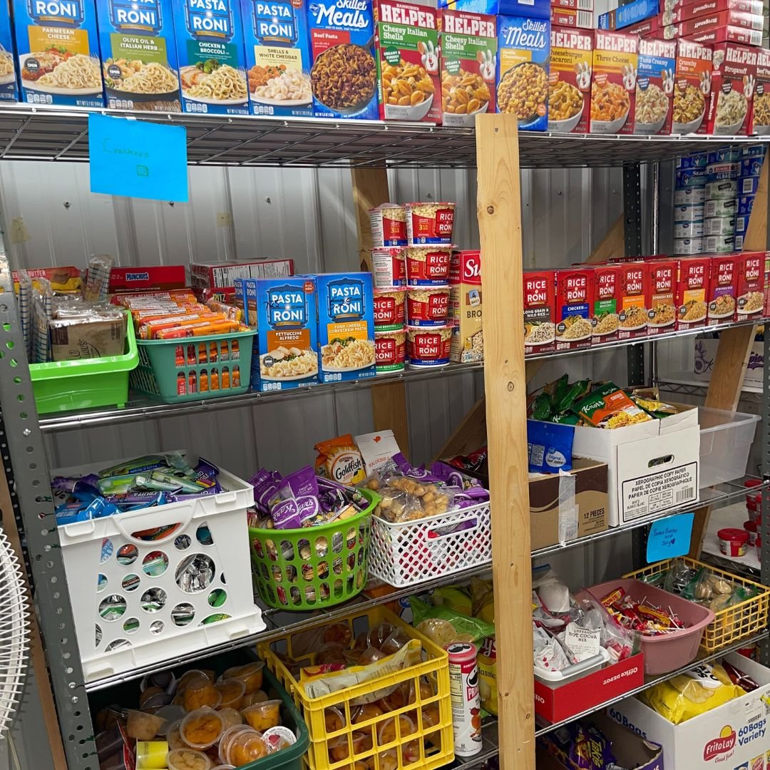 A grocery store shelf stocked with various food items including canned pasta, boxed macaroni and cheese, ramen noodles, snack packs, and other packaged snacks.
