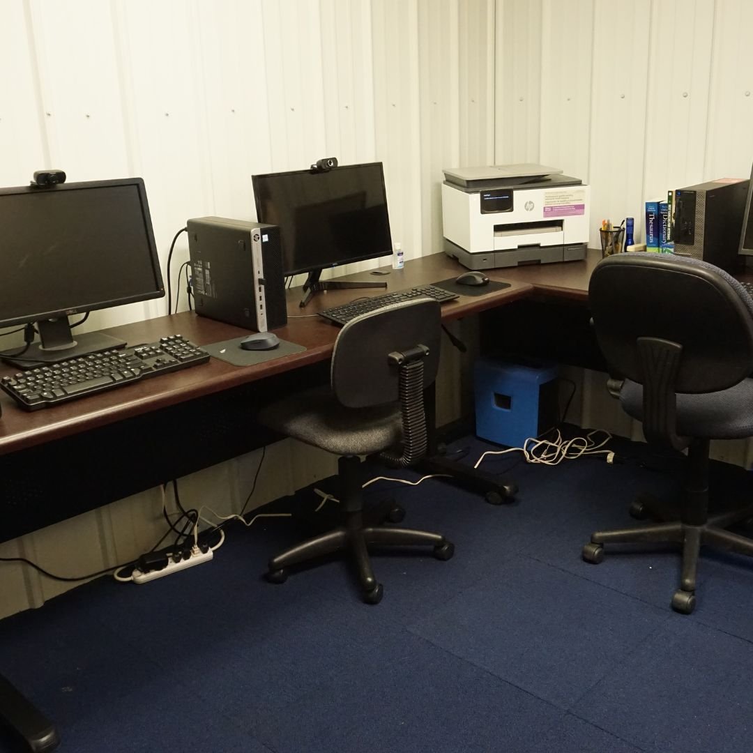 An office workspace with two computer desks, each with a monitor, keyboard, and mouse. There is a printer on the right desk and a small shelving unit with books. The floor is carpeted in dark blue, and there are two office chairs with wheels.