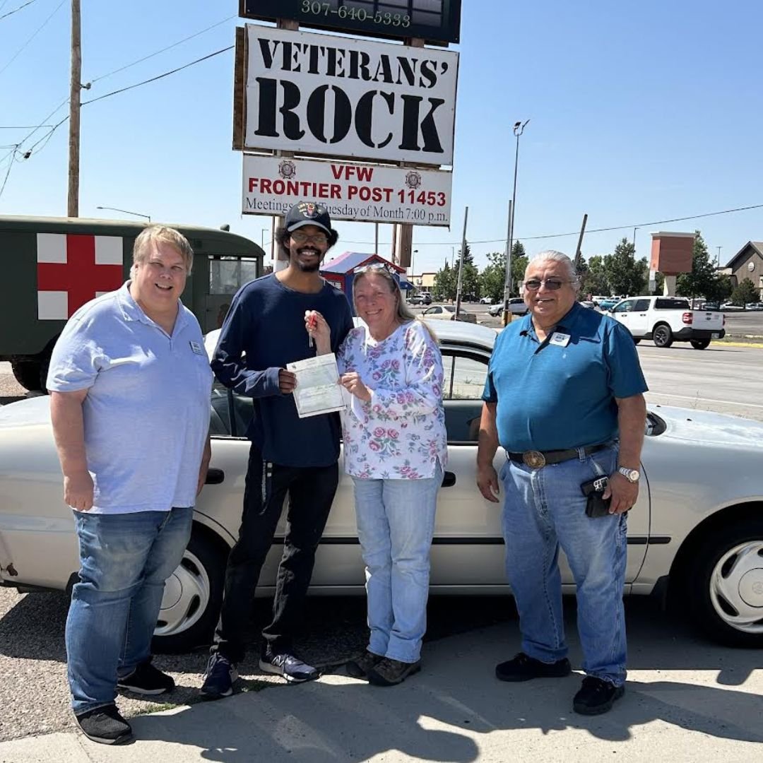 Four people standing in front of a car, smiling, during a Veterans' Rock event at VFW Frontier Post 11453, with a sign in the background.