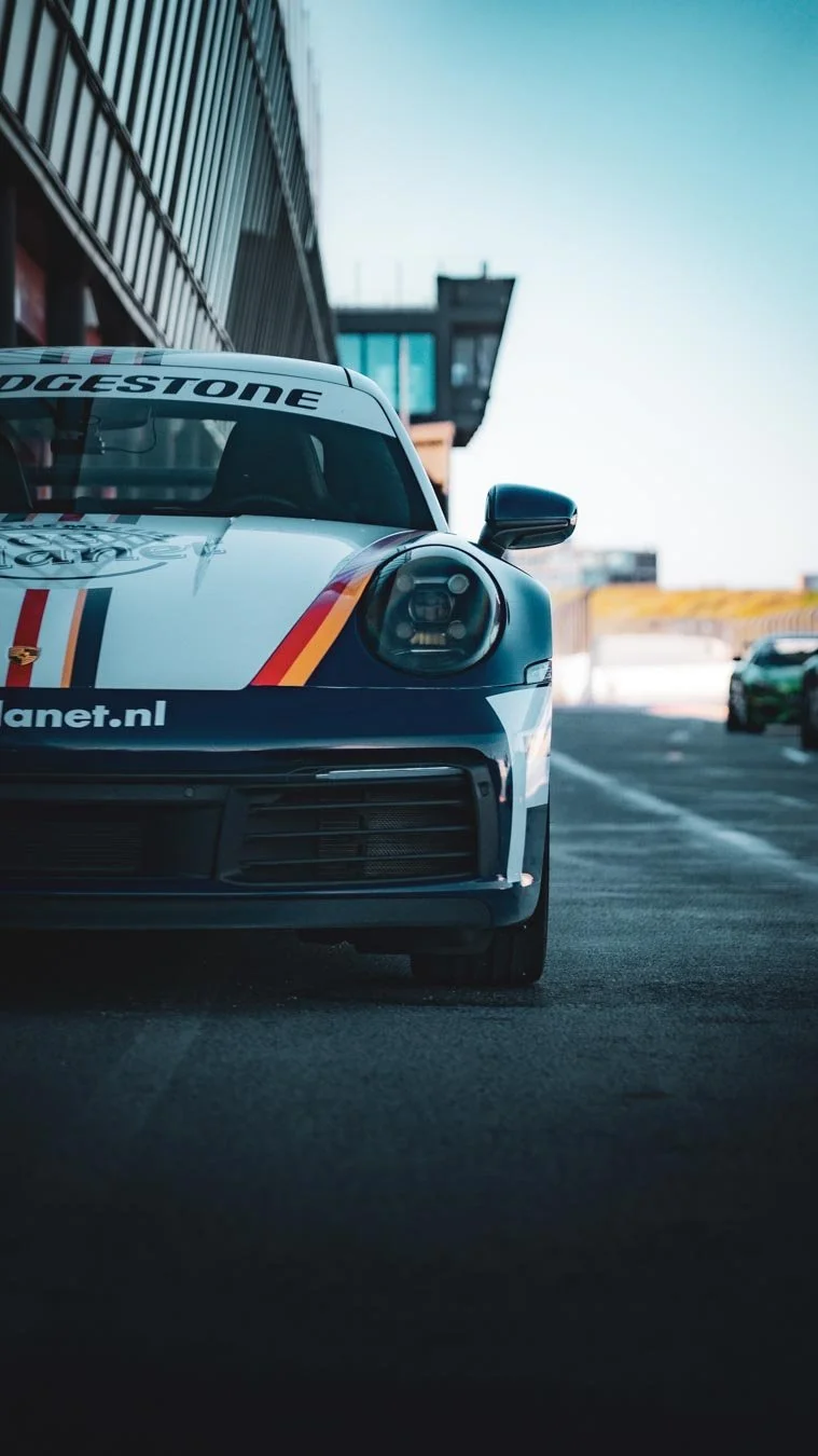 Ein Porsche Rennwagen im Boxengassenbereich der Rennstrecke mit Bridgestone und anderen Sponsorenlogos auf dem Auto. Das Auto steht neben einer modernen Gebäudekonstruktion, die zu einem Rennstadion gehört.