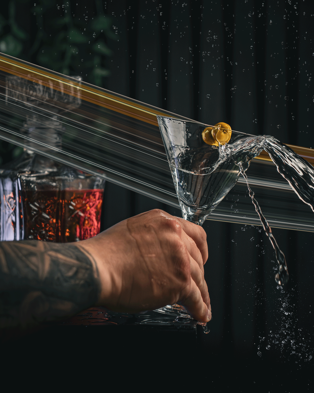 Ein Hand mit Tattoos hält ein Martini-Glas, während Wasser über das Glas läuft, mit einem dunklen Hintergrund und einer im Hintergrund.