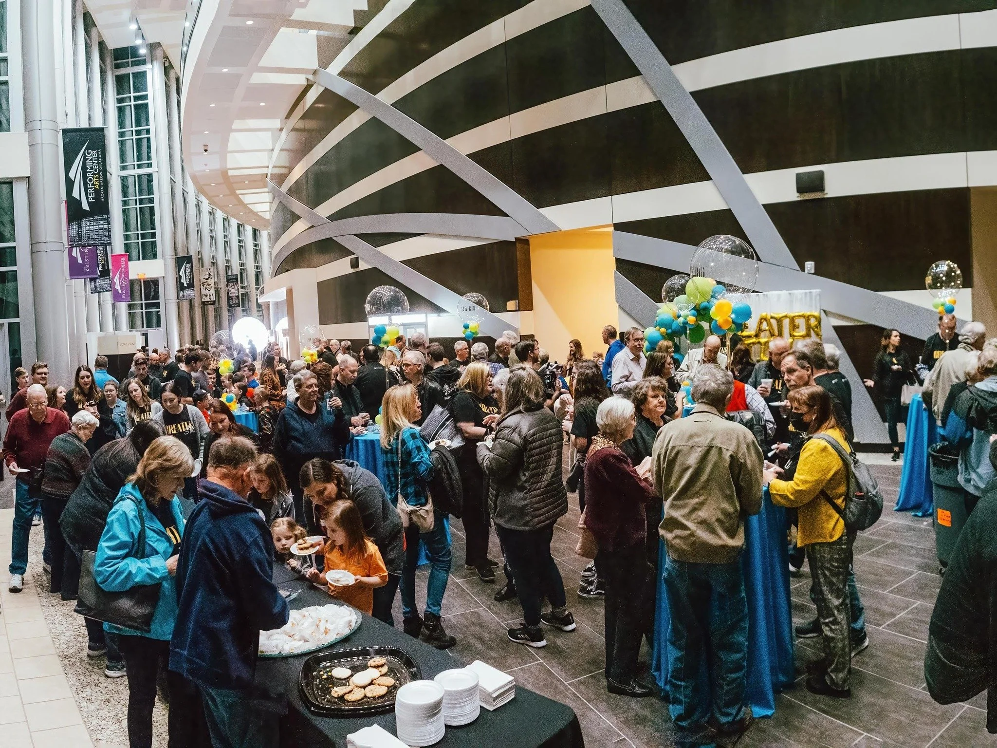 Crowd of people at an indoor event with balloon decorations and a banner that says 'ATLR' in the background. Food served at tables in the foreground.