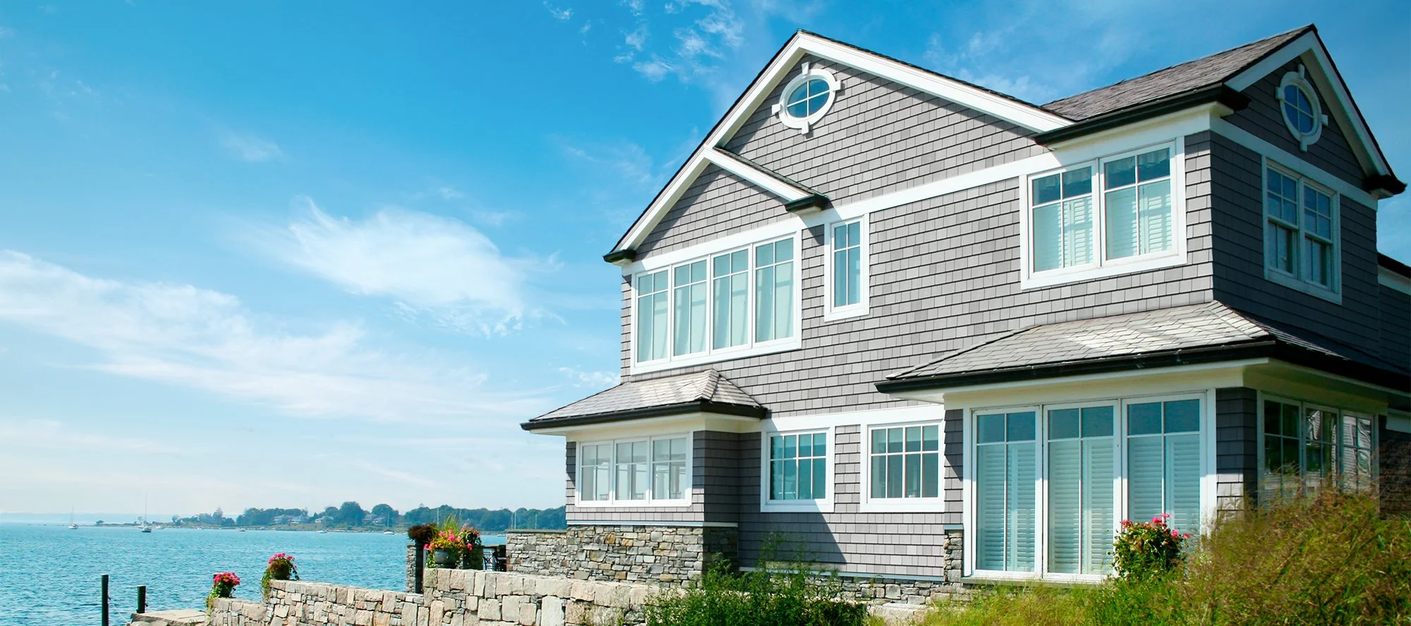 A large, multi-story house with gray shingle siding and multiple windows, situated by a body of water under a clear blue sky.