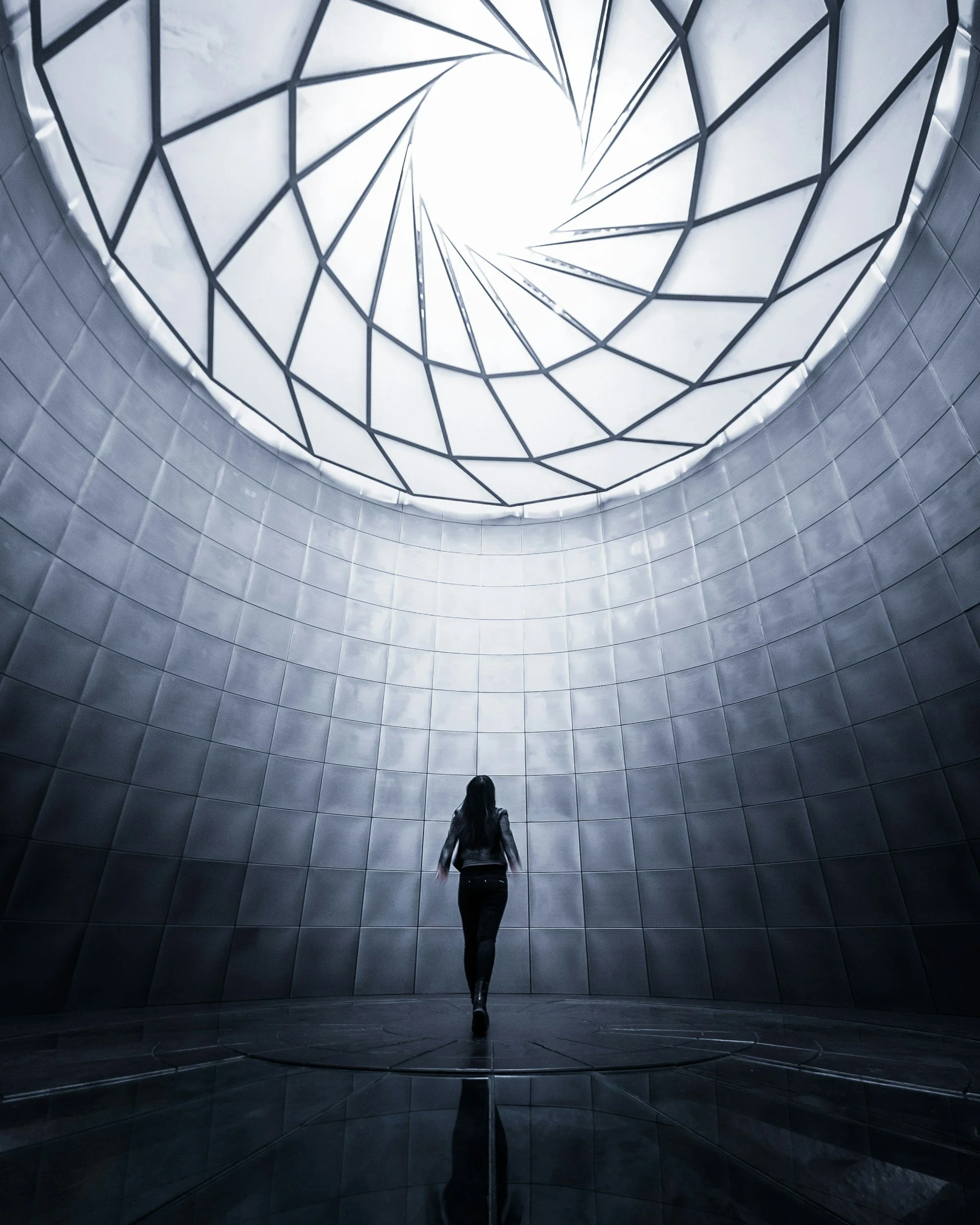 A woman in dark clothing walking inside a circular, modern architectural space with tiled walls and a geometric glass ceiling that allows natural light to enter.