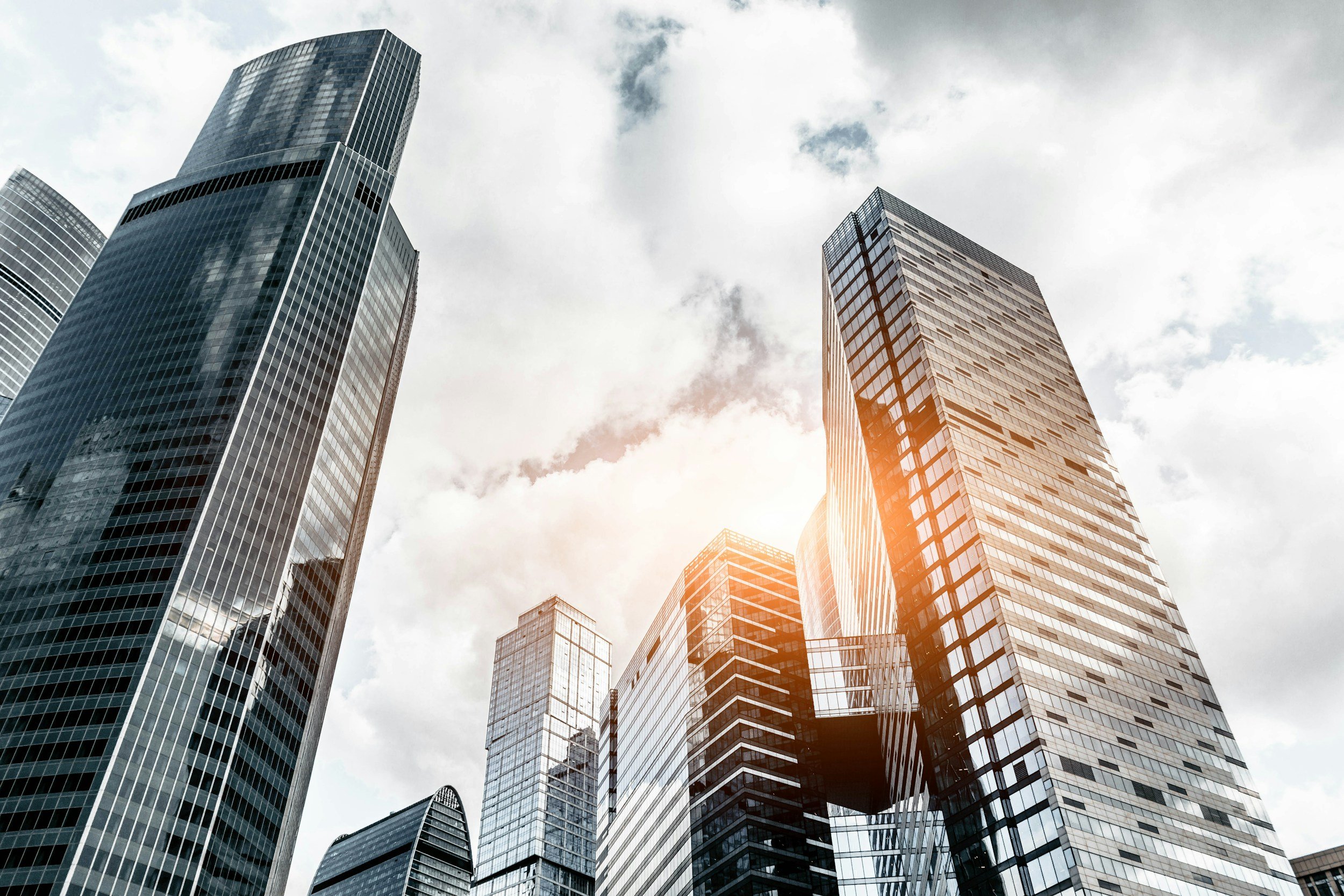A cluster of tall modern skyscrapers in a city with cloudy sky and sunlight reflecting off the glass buildings.