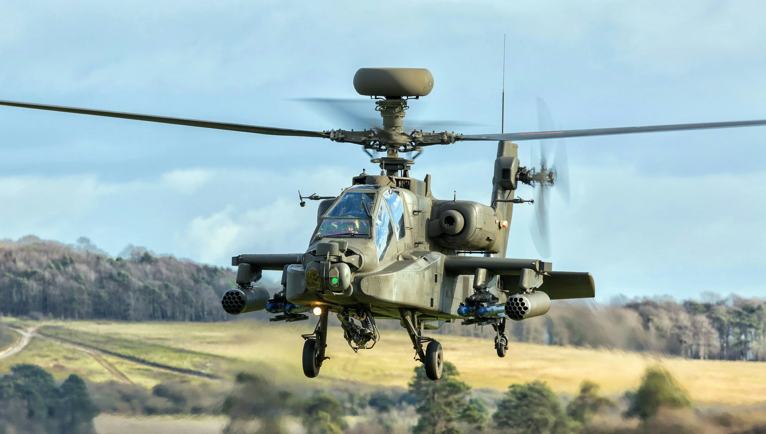 Military attack helicopter flying over a rural landscape with fields and trees.