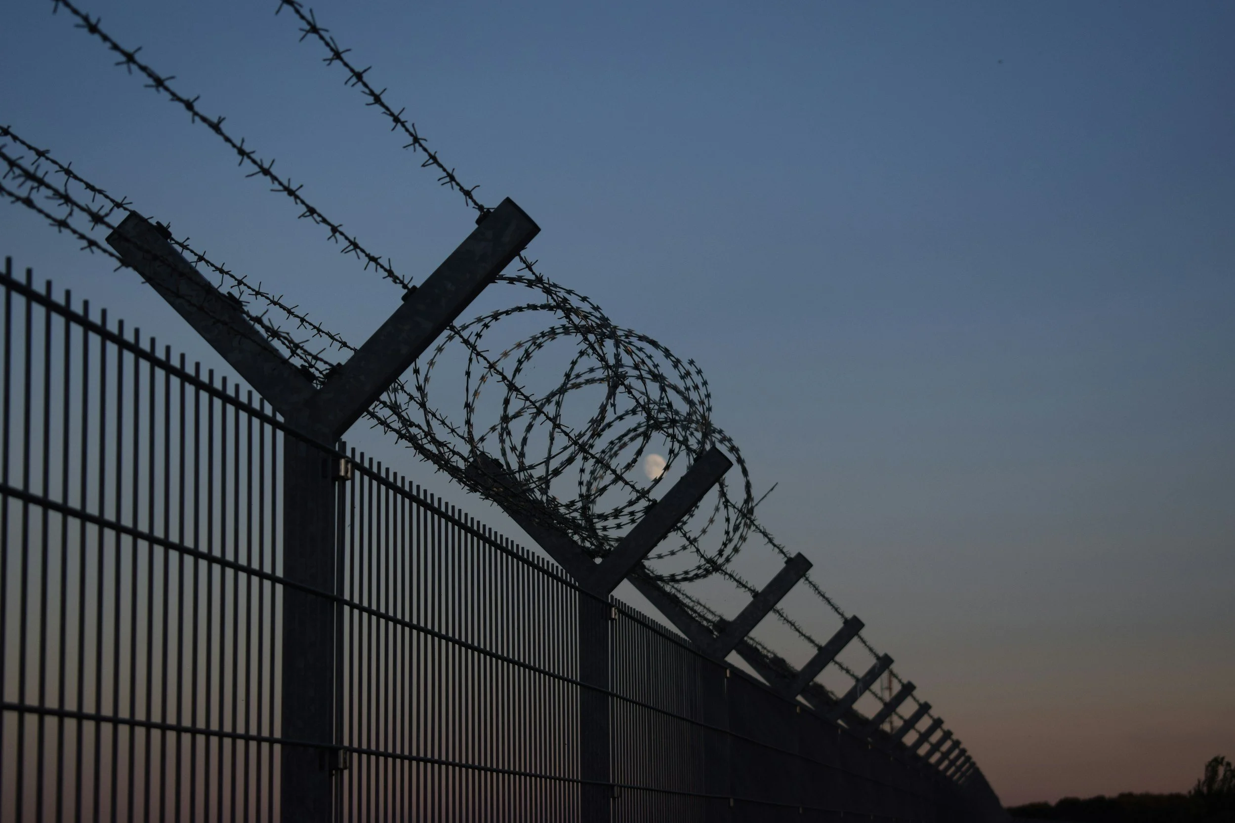 A tall metal security fence with barbed wire coils along the top, set against a dusk sky with a visible moon.