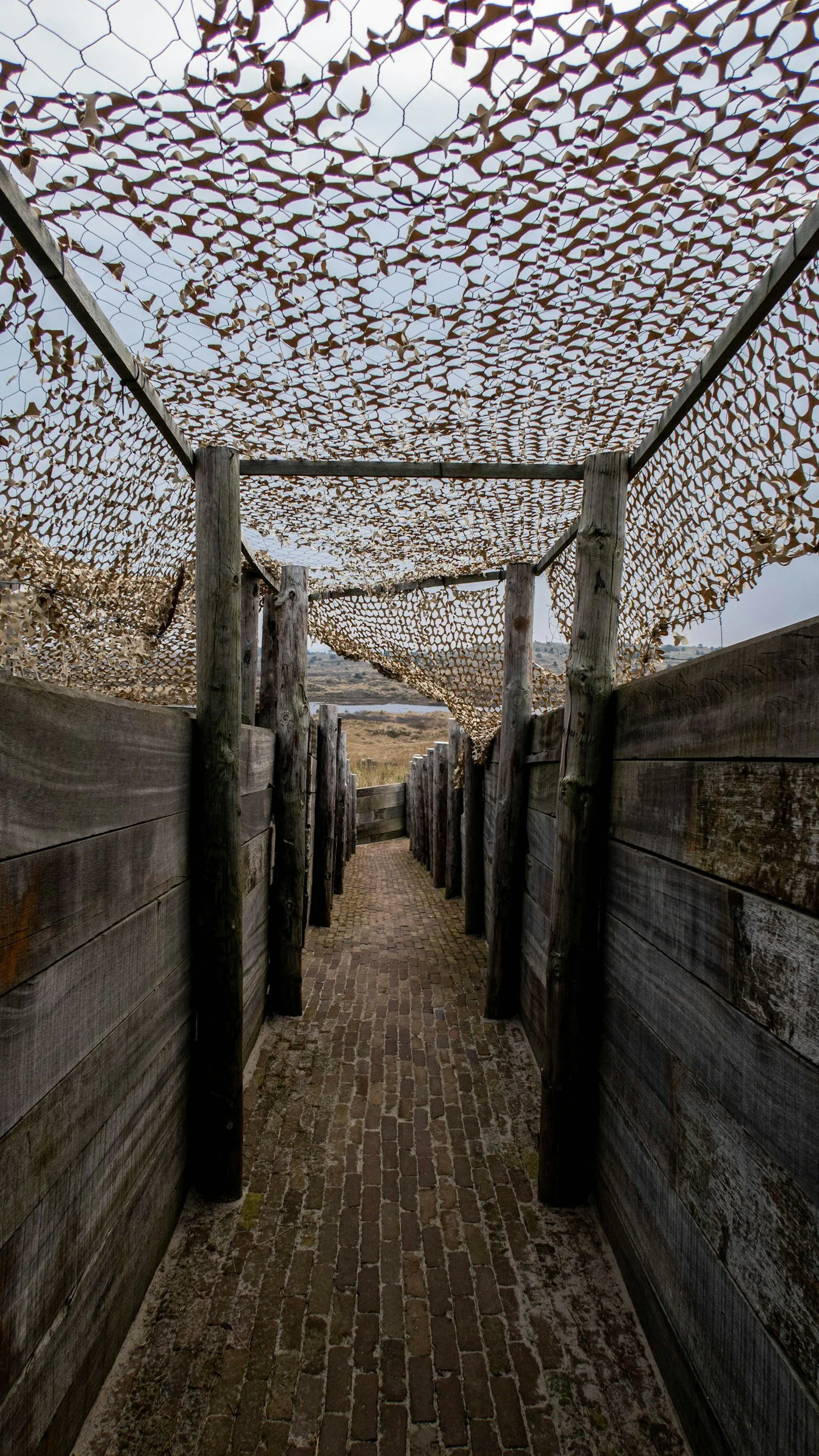 Narrow brick pathway surrounded by wooden walls with camouflage netting overhead.