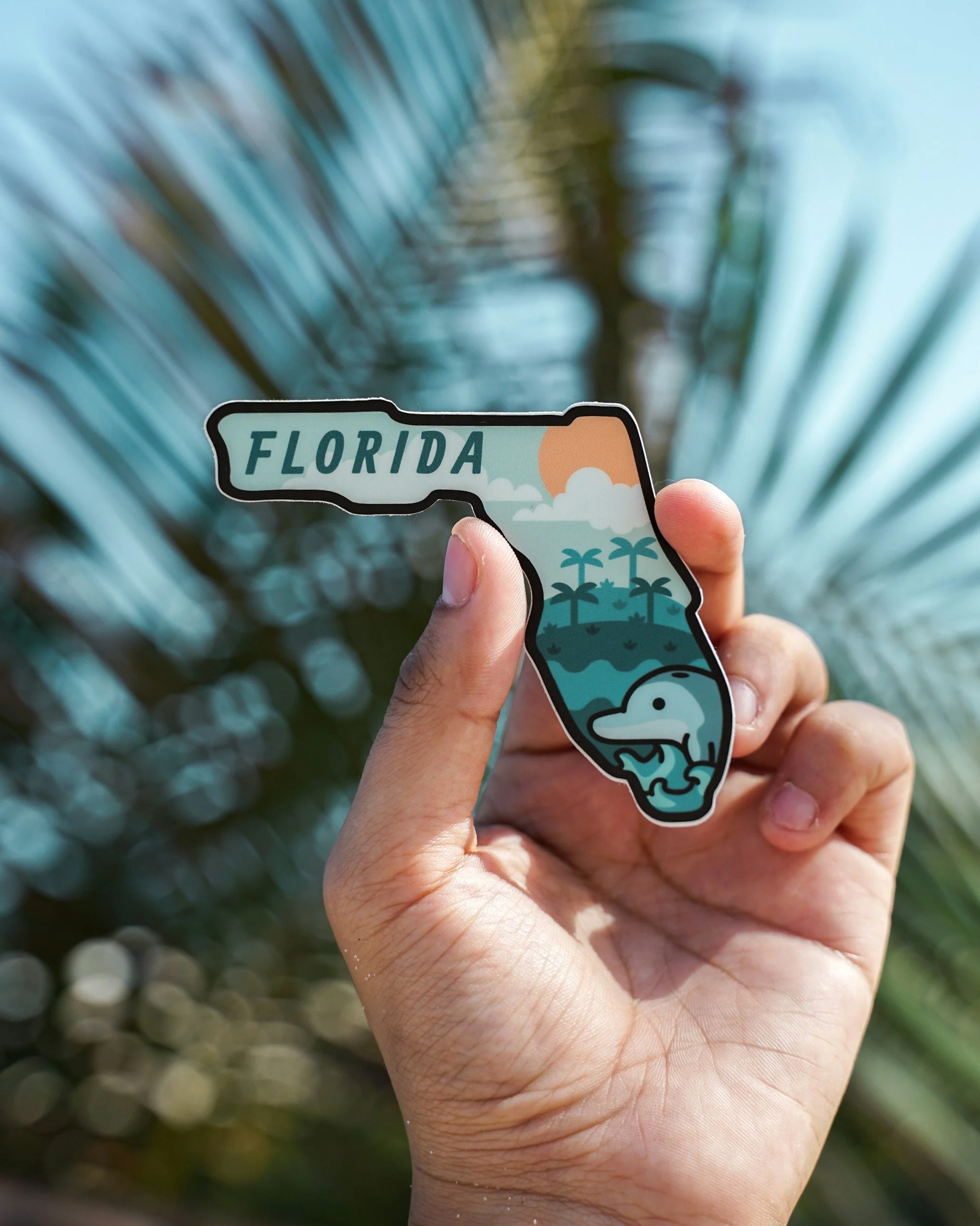 A hand holds a sticker shaped like the state of Florida with illustrations of palm trees, a sunset, clouds, waves, and a dinosaur inside, with the word 'Florida' on it. In the background, blurred palm tree leaves are visible.