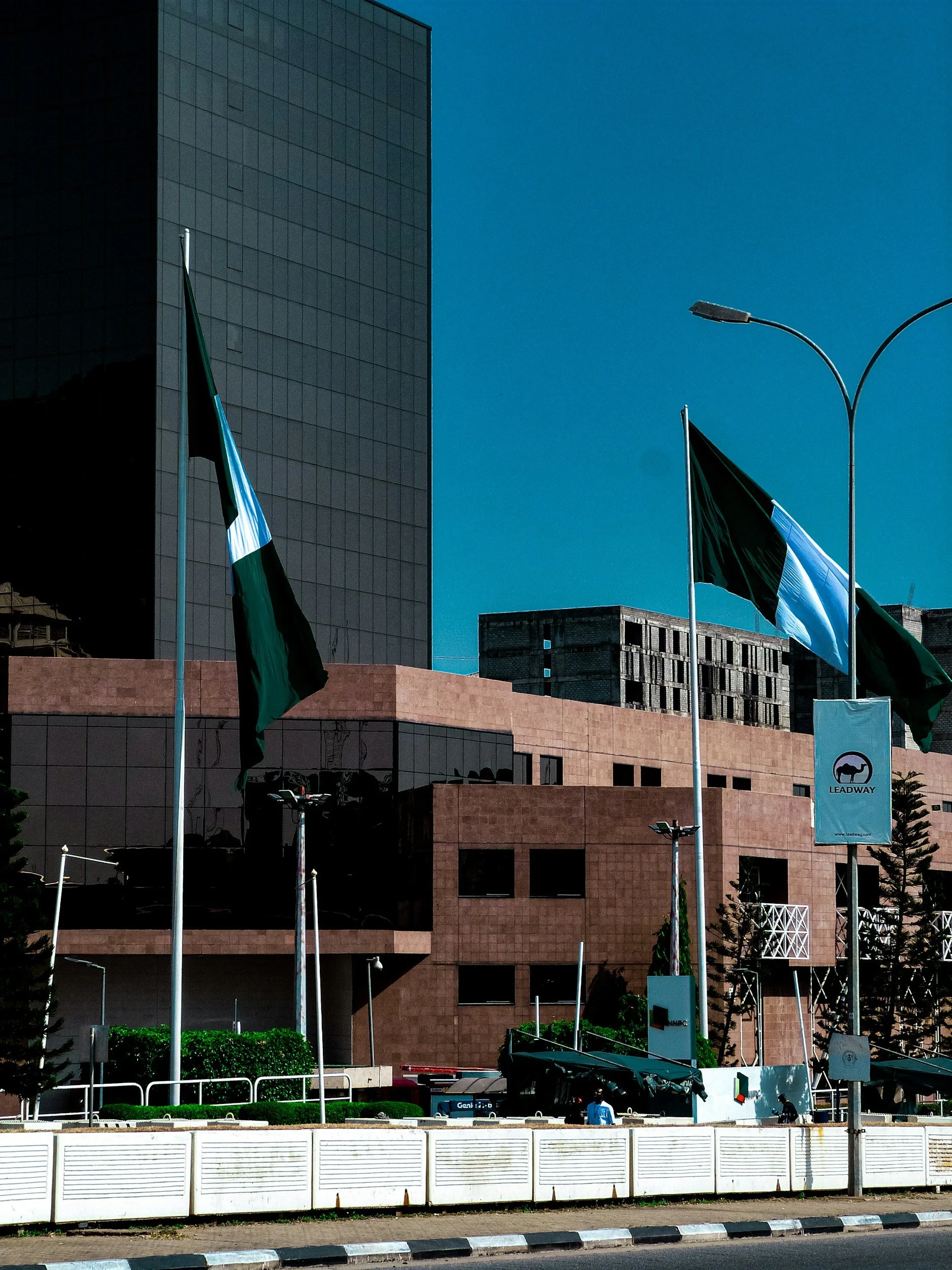 Urban scene with modern glass office building, flagpoles with black and green flags, streetlights, and a banner with a logo and the word 'Leadway' in front of a beige brick structure.
