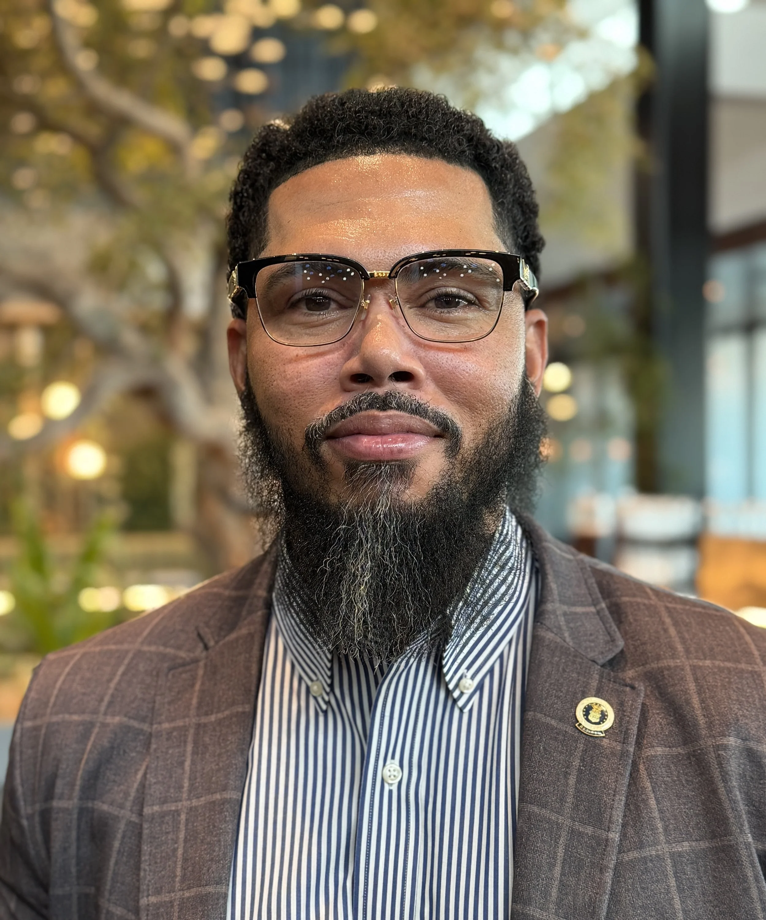 A man with glasses, a beard, and a mustache wearing a brown checkered blazer and a striped dress shirt, standing outdoors with trees and a building in the background.