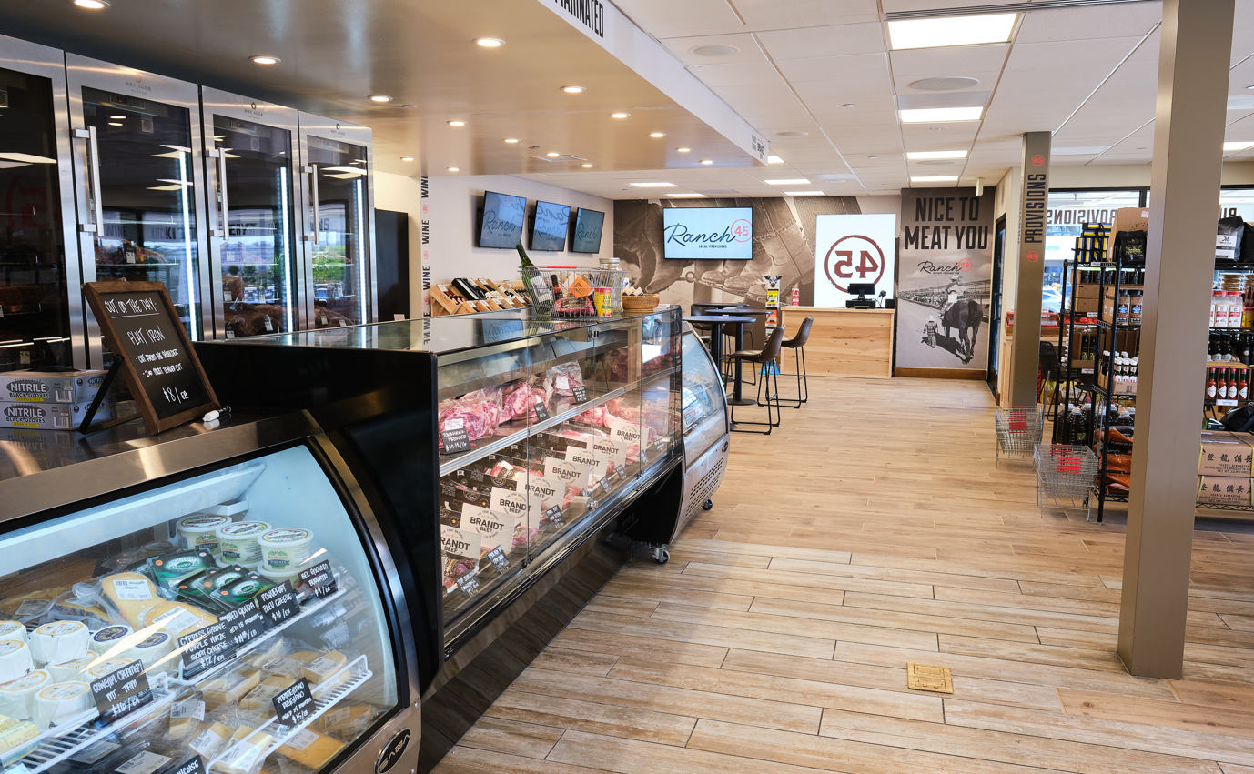 Inside a deli or grocery store with a glass display case of meats and cheeses, a refrigerated section with dairy products, a small seating area with tables and chairs, and a wall with signs including 'Nice to Meat You' and 'Provision'.
