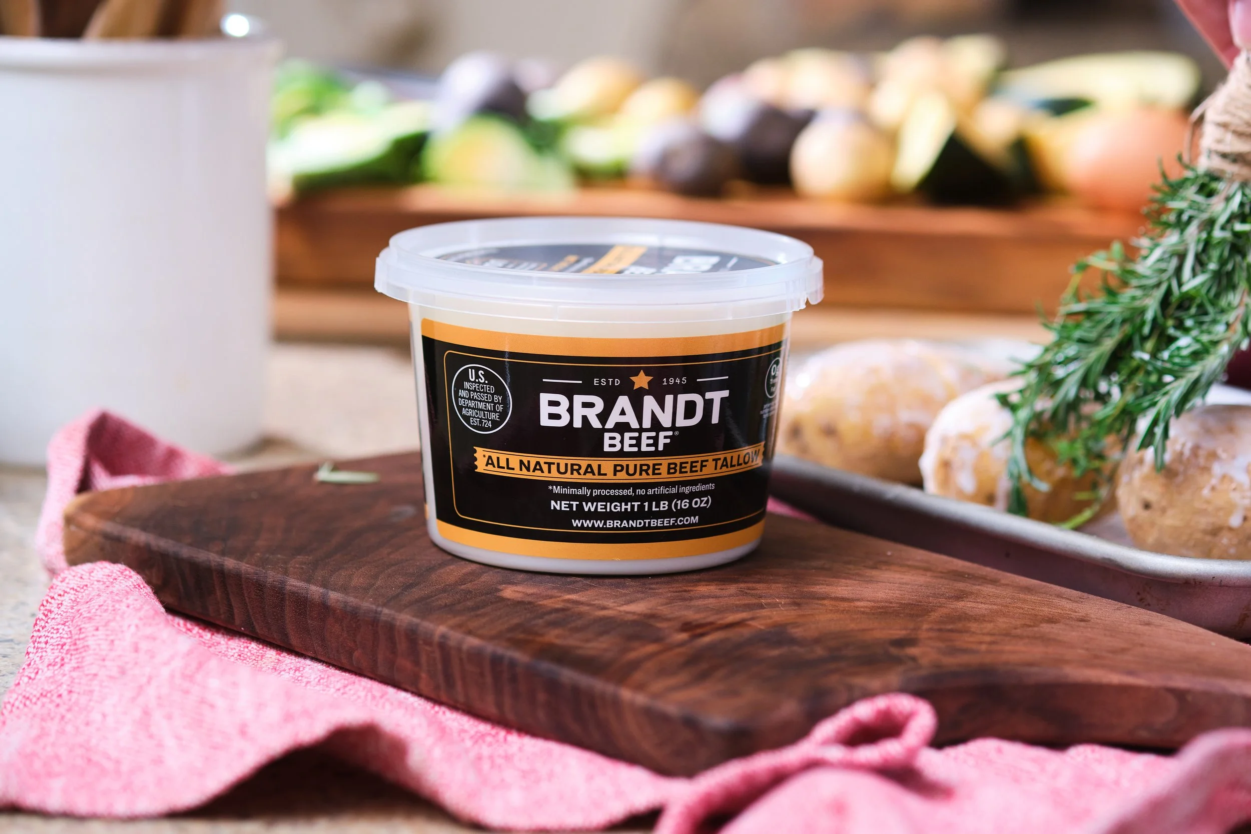 Container of Brandt Beef all natural pure beef tallow placed on a wooden cutting board with a pink cloth, with baked goods and herbs in the background.