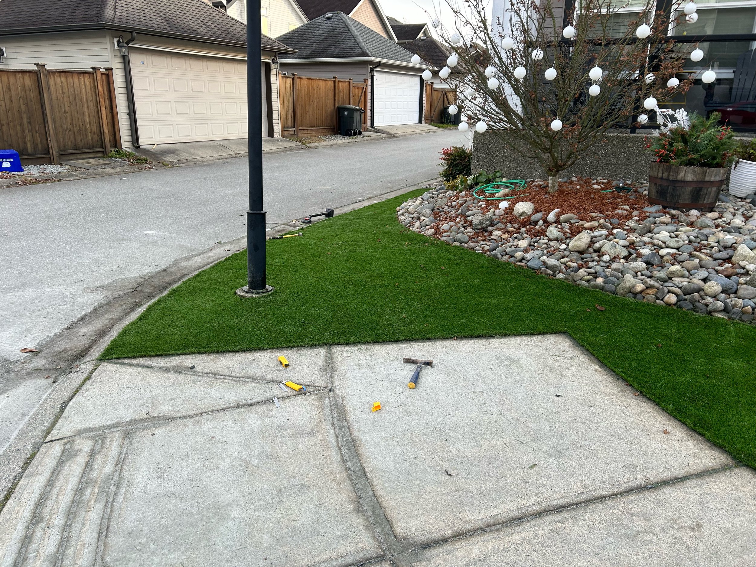 Suburban alleyway with artificial grass, a streetlamp, tree with hanging lights, rocks, and garden tools on pavement.