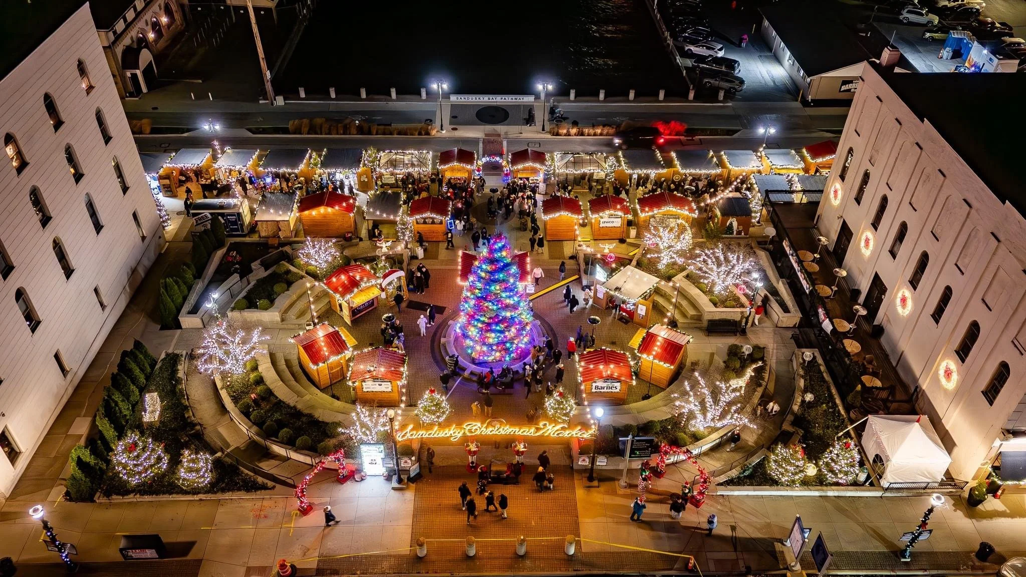 Aerial view of a Christmas market at night with lit decorations, a large lit Christmas tree, and festive huts, surrounded by buildings and parked cars.