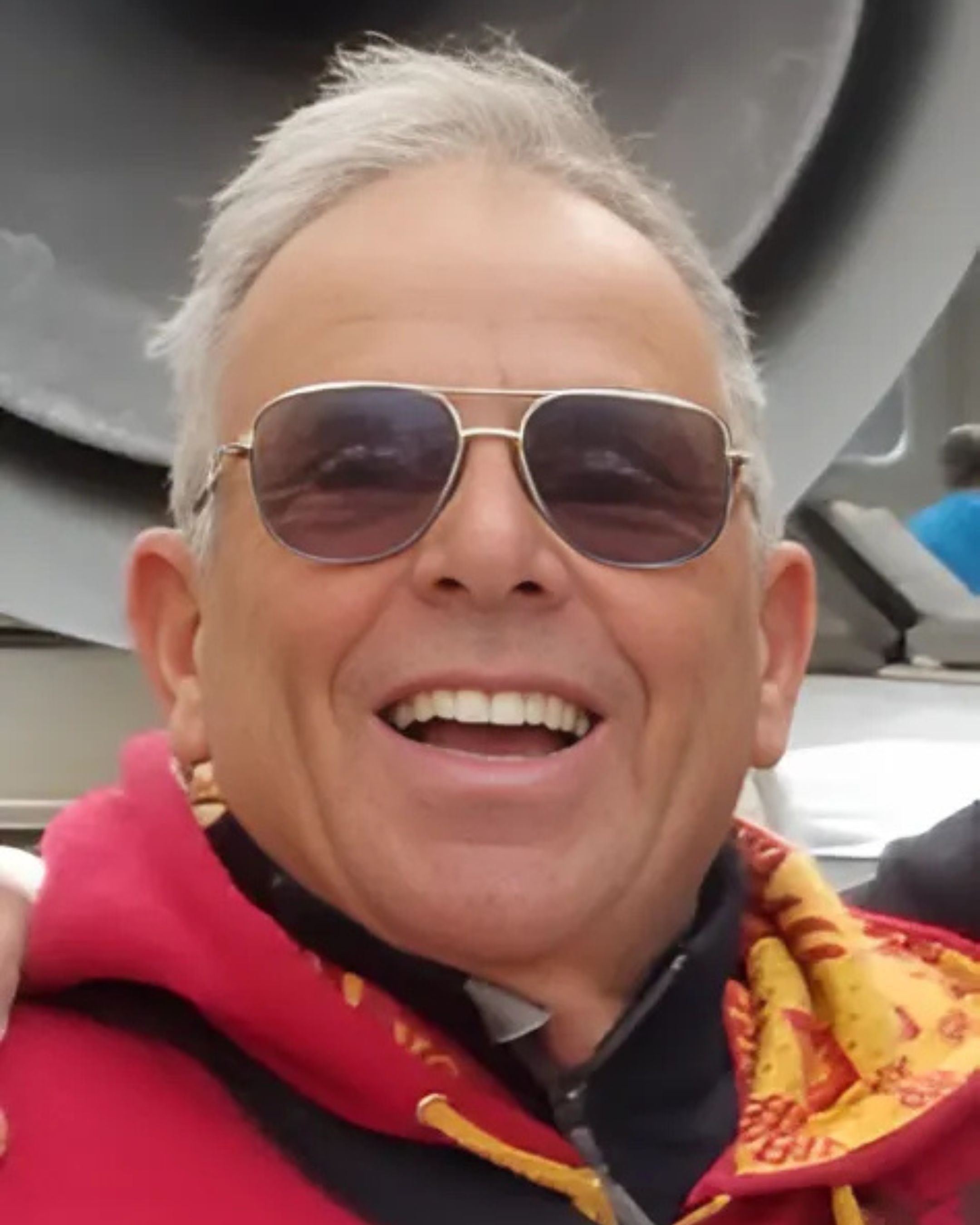 Close-up of a smiling person wearing sunglasses, with white or gray hair, in an indoor setting.