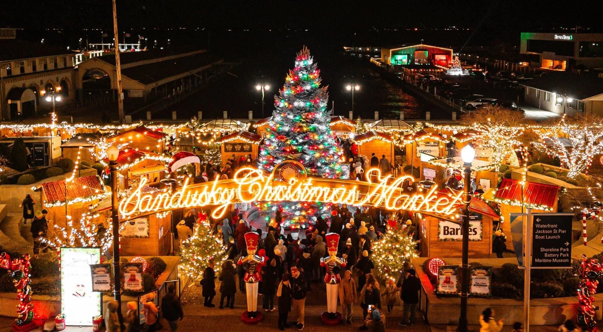 Nighttime view of the Sandusky Christmas Market decorated with Christmas lights, a large decorated Christmas tree, and festive wooden stalls. The market is crowded with people and there's a sign with glowing orange script that reads 'Sandusky Christmas Market.' In the background, there are buildings and parking lots illuminated, with some Christmas decorations visible.
