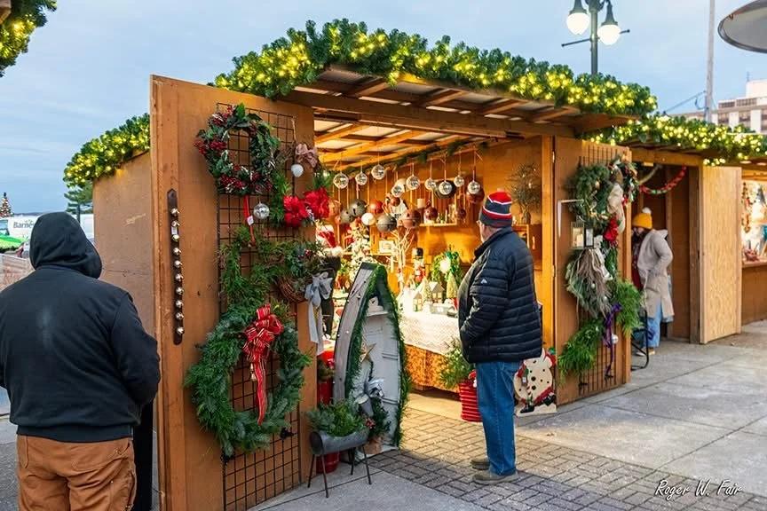 People browsing a Christmas market stall decorated with holiday wreaths, ornaments, and greenery.