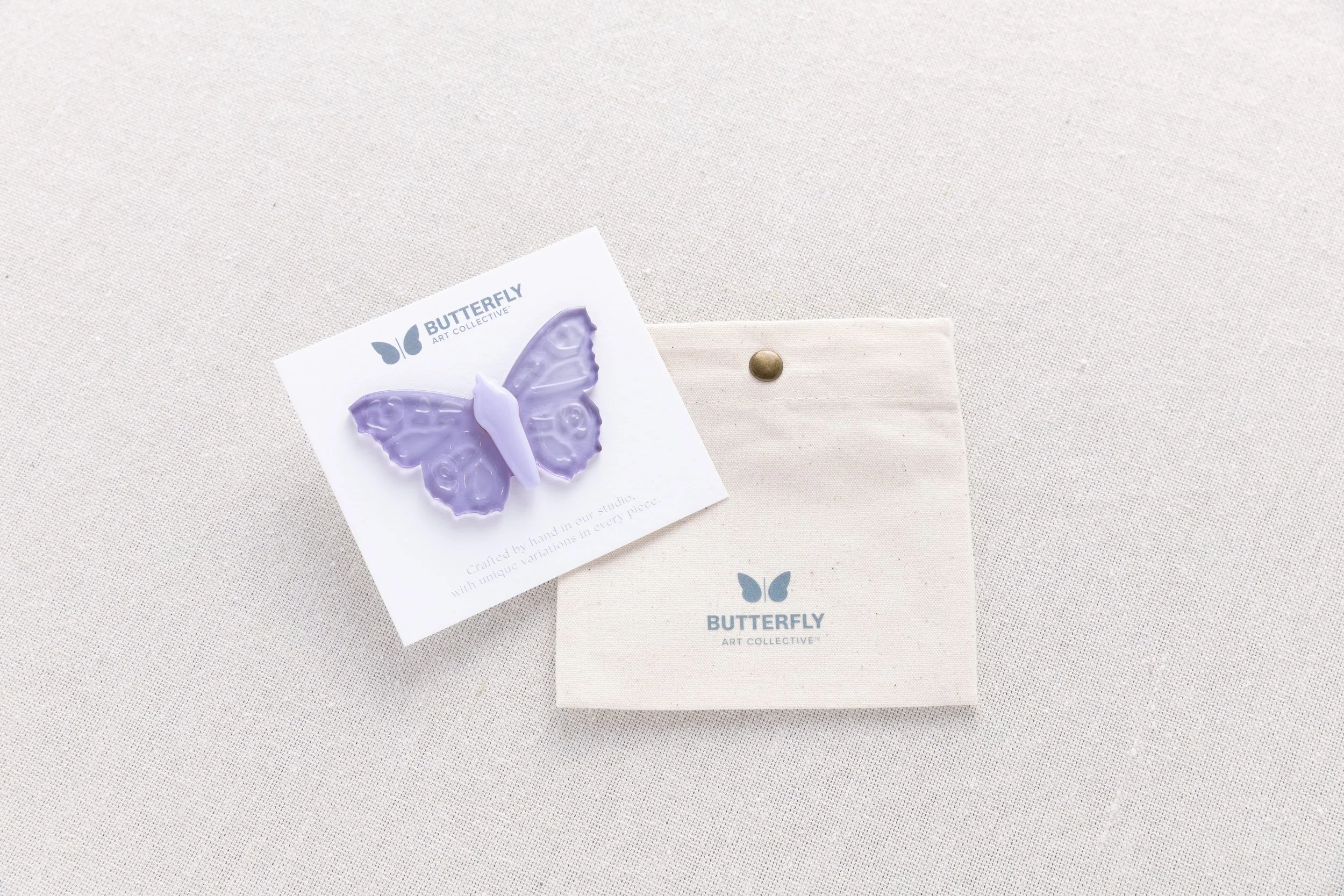 Twilight Glass soft resin butterfly pin in translucent lavender with a lilac body, displayed on a branded card with a cotton canvas pouch.