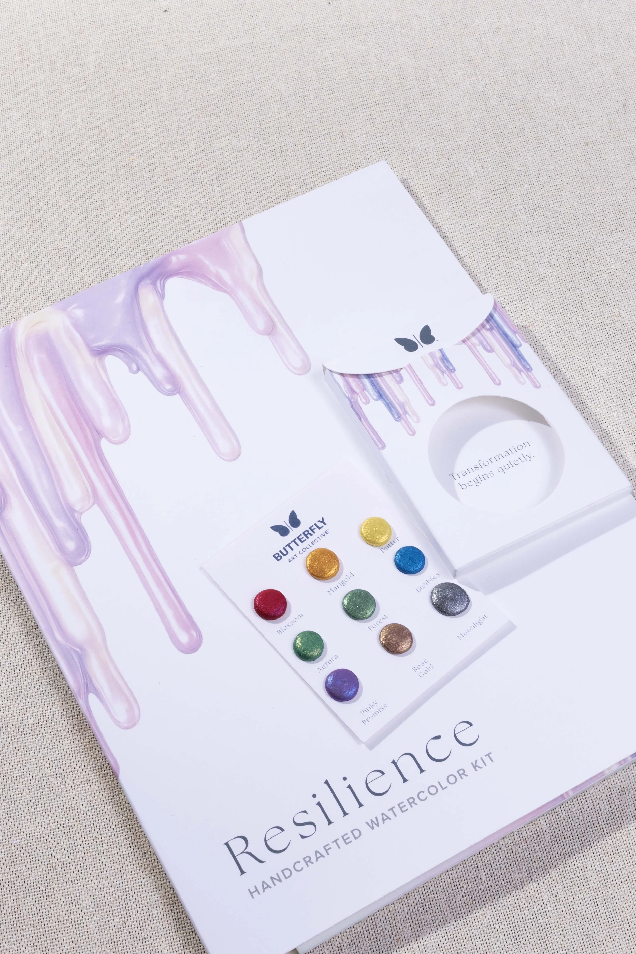 Overhead view of the Resilience watercolor kit packaging featuring lavender paint drips, the watercolor palette card, and product components.