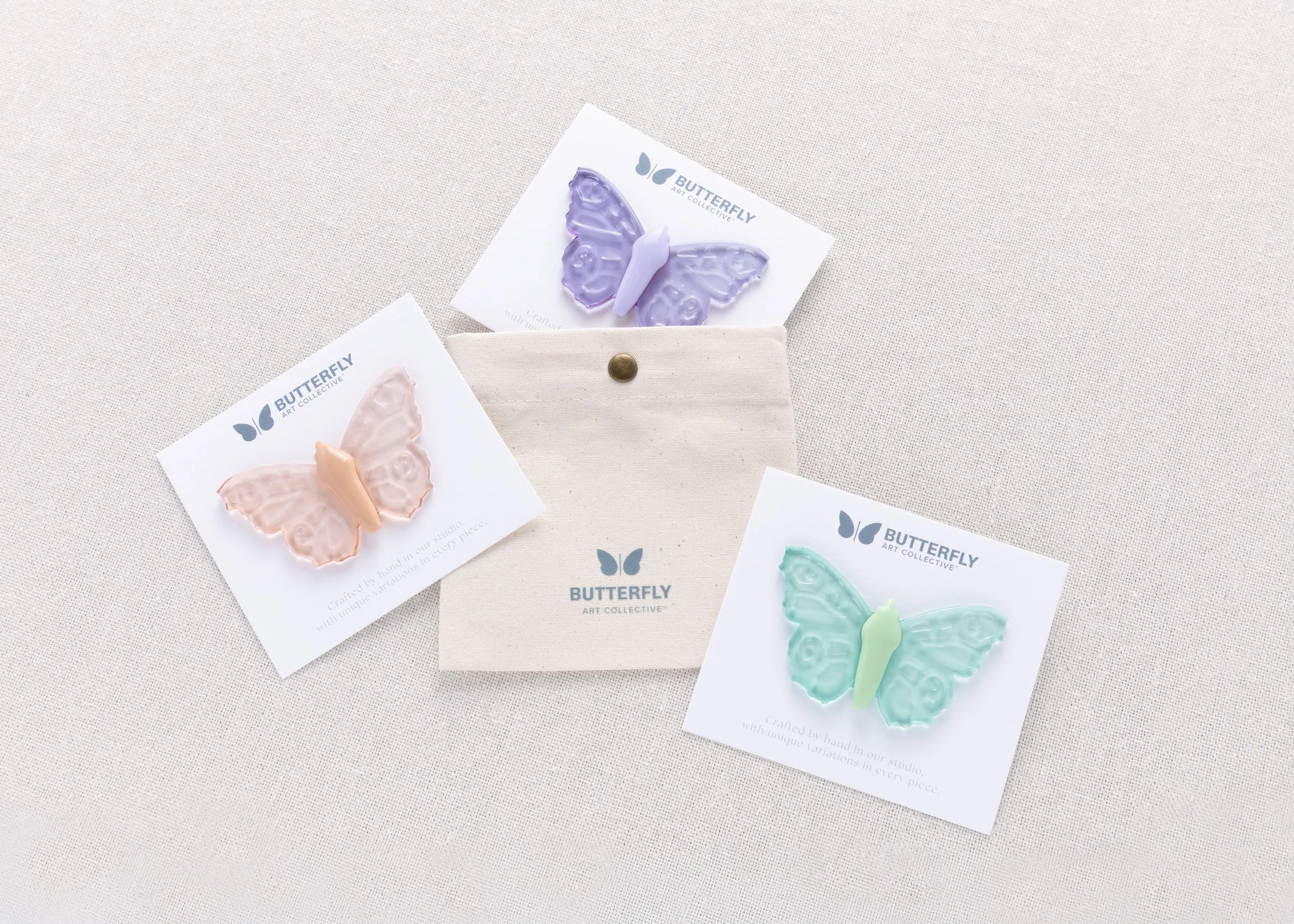Butterfly Art Collective soft resin butterfly pins in three translucent colors, presented on individual display cards alongside a branded canvas pouch.