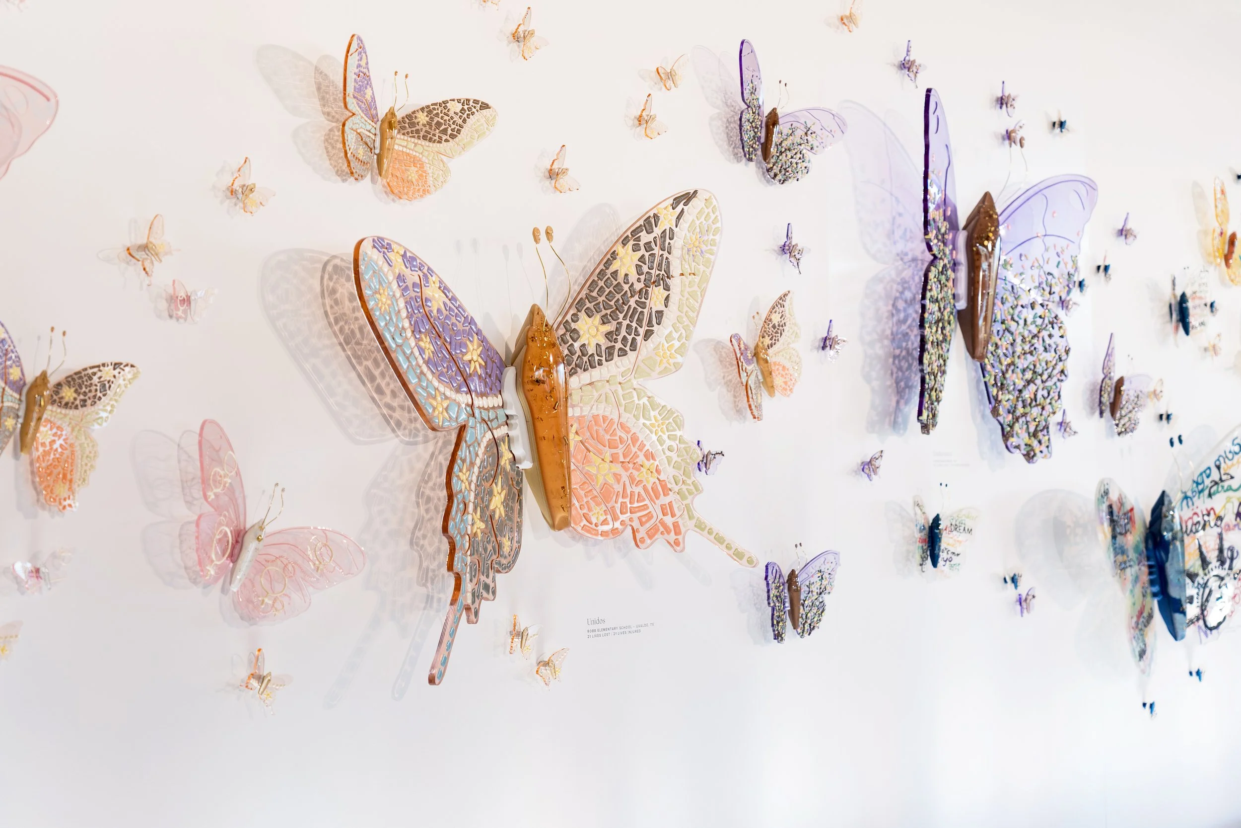 Wall display of colorful resin butterfly sculptures with various intricate patterns and shadows, arranged on a white wall.